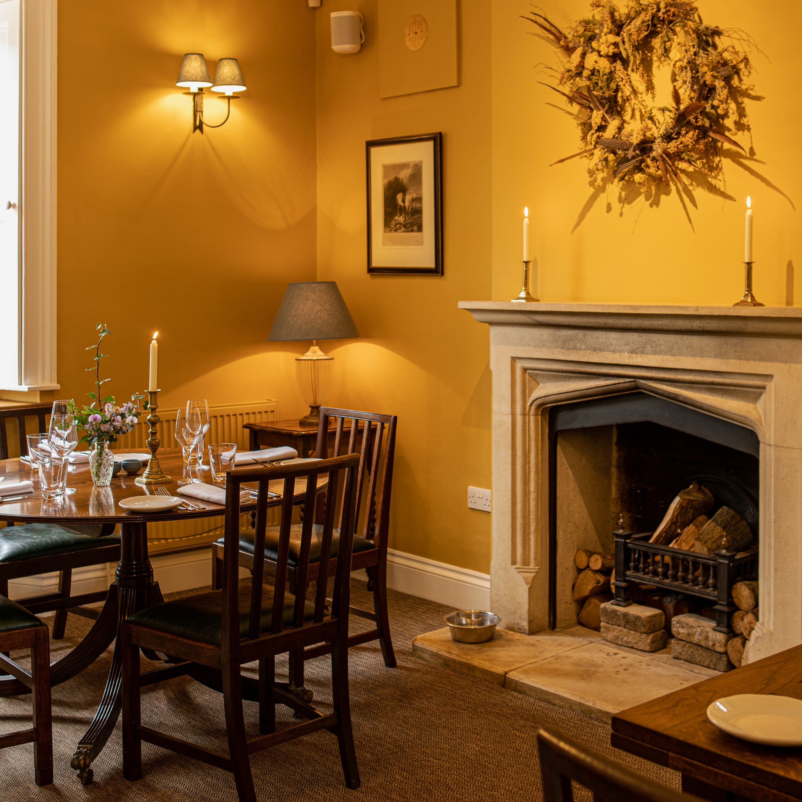 Cozy restaurant dining area with wooden table and chairs, set for a meal, next to a stone fireplace with candles and a wreath above it.