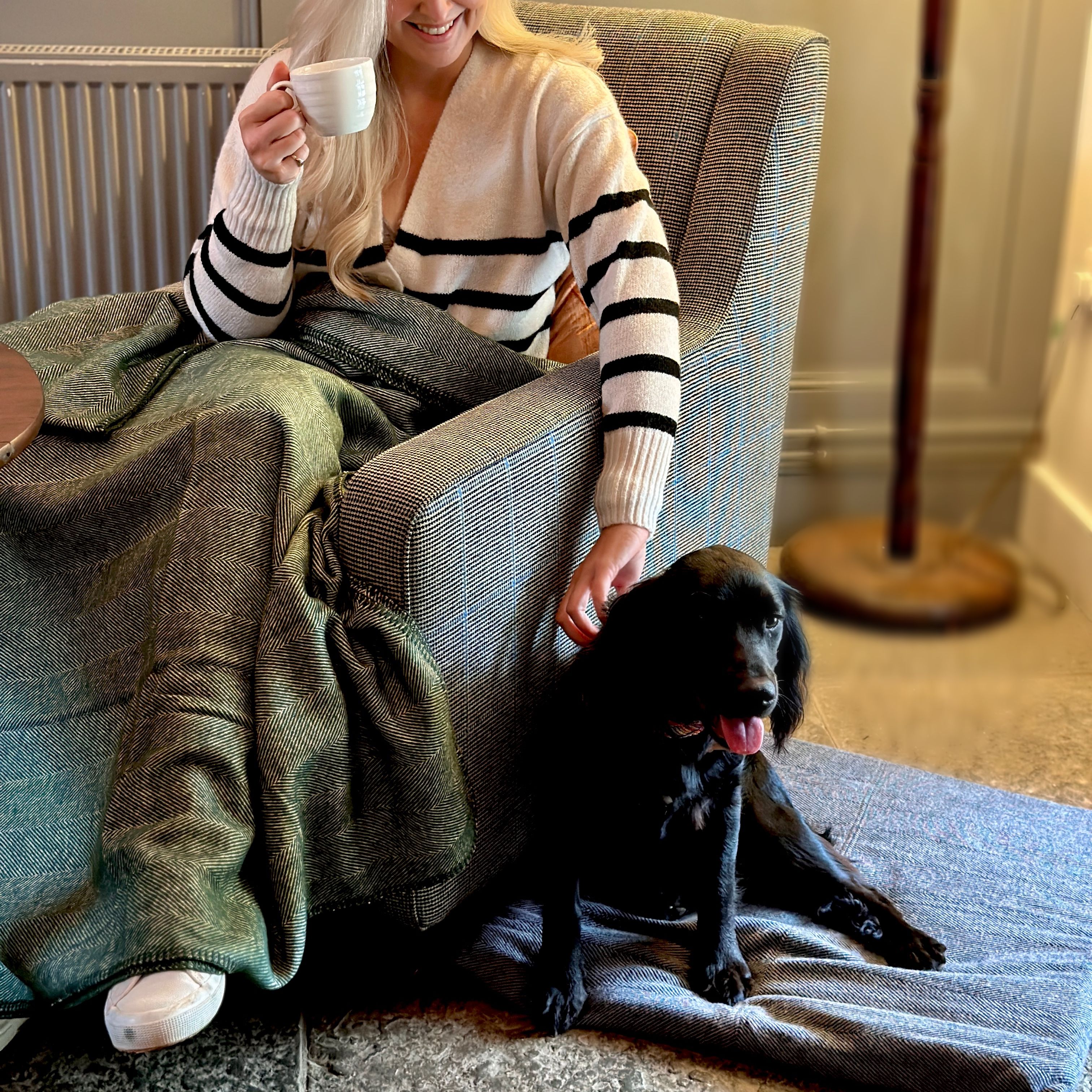 Woman sitting in an armchair with a blanket, holding a cup, and petting a black dog sitting on a mat beside her.