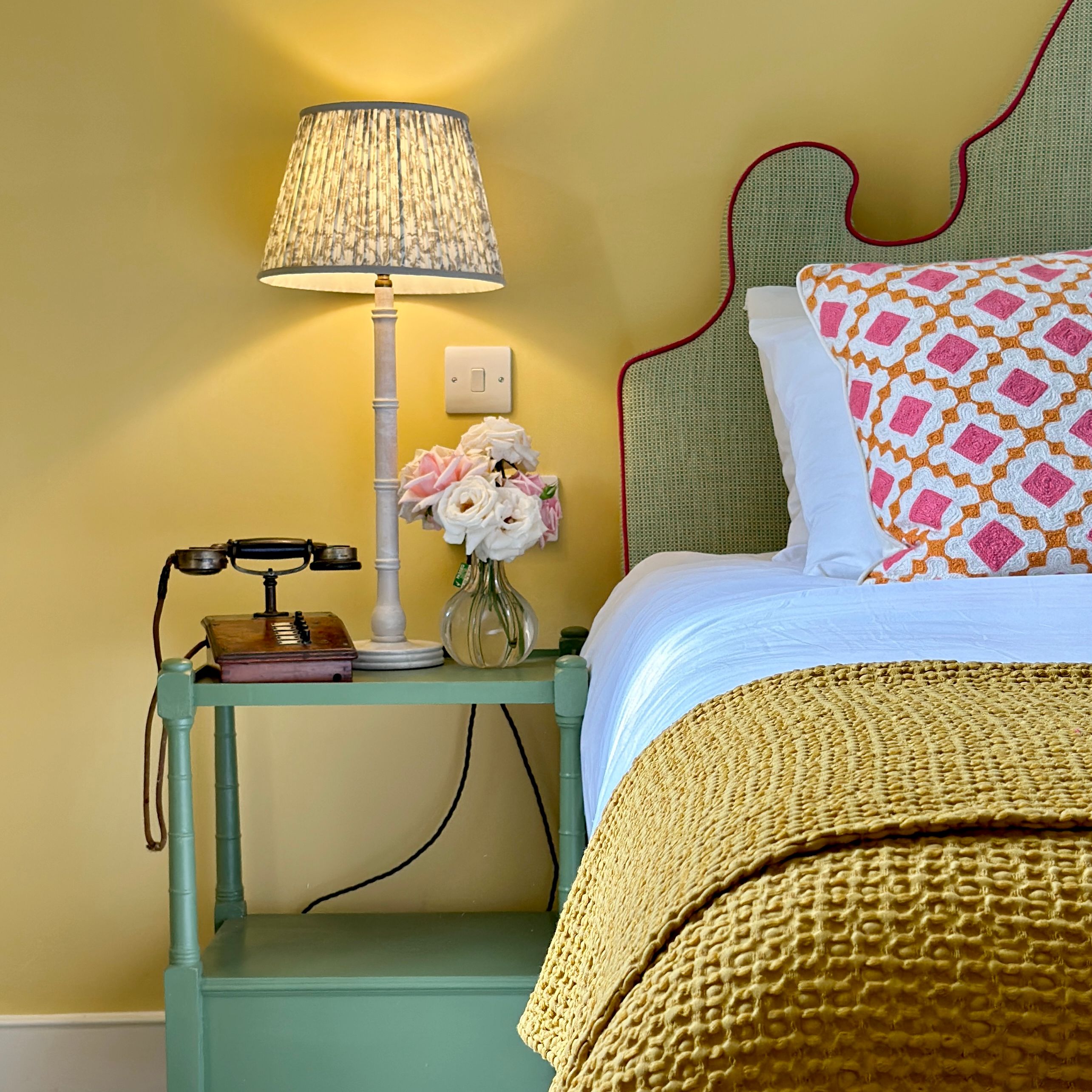 Cozy bedroom scene with a green nightstand, vintage telephone, table lamp, vase of flowers, and slippers on the floor.
