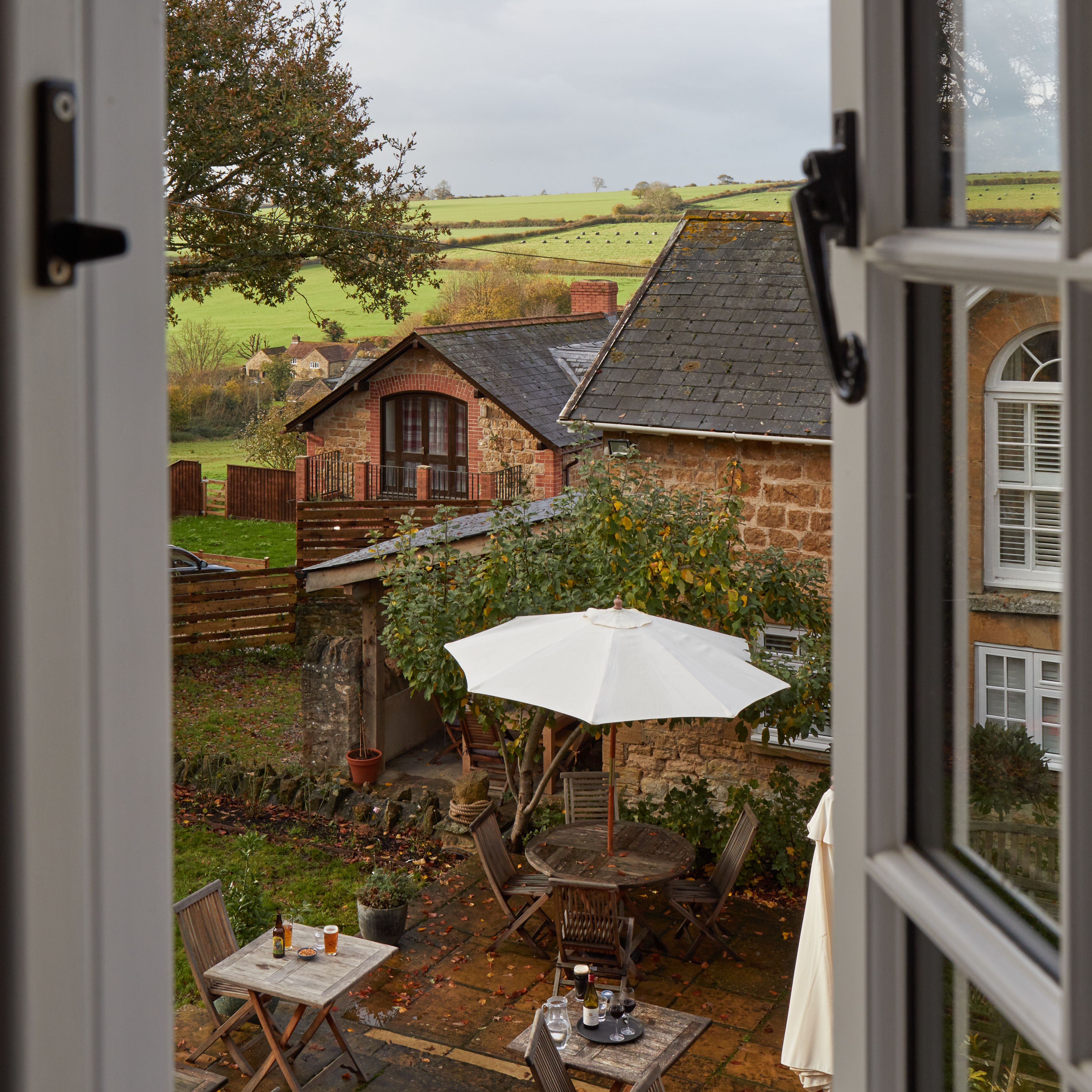 A view through an open window overlooking a countryside patio with wooden tables, chairs, and a white umbrella.