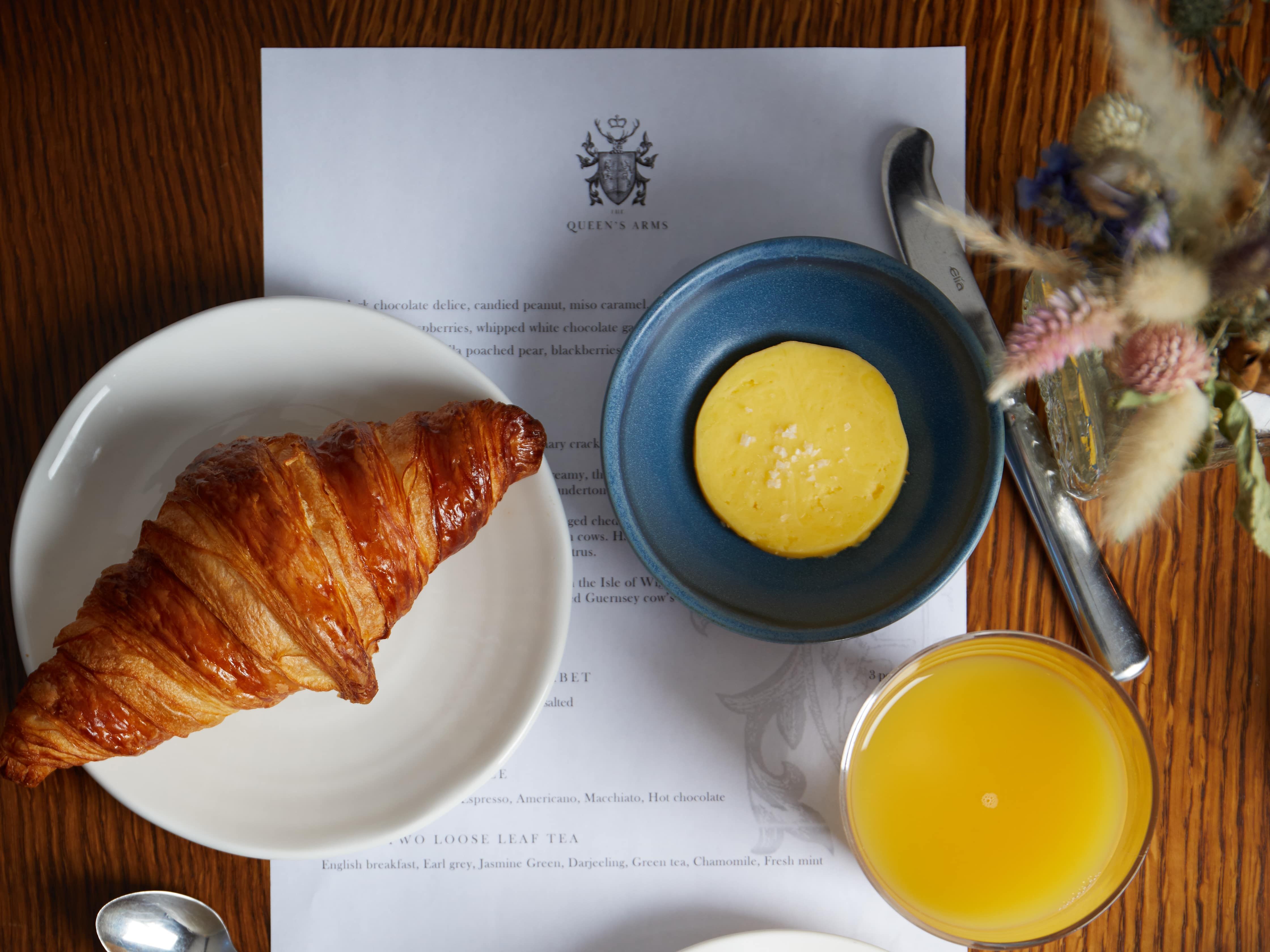 Breakfast table with croissant, butter, orange juice, cappuccino, apples, granola, and a teapot