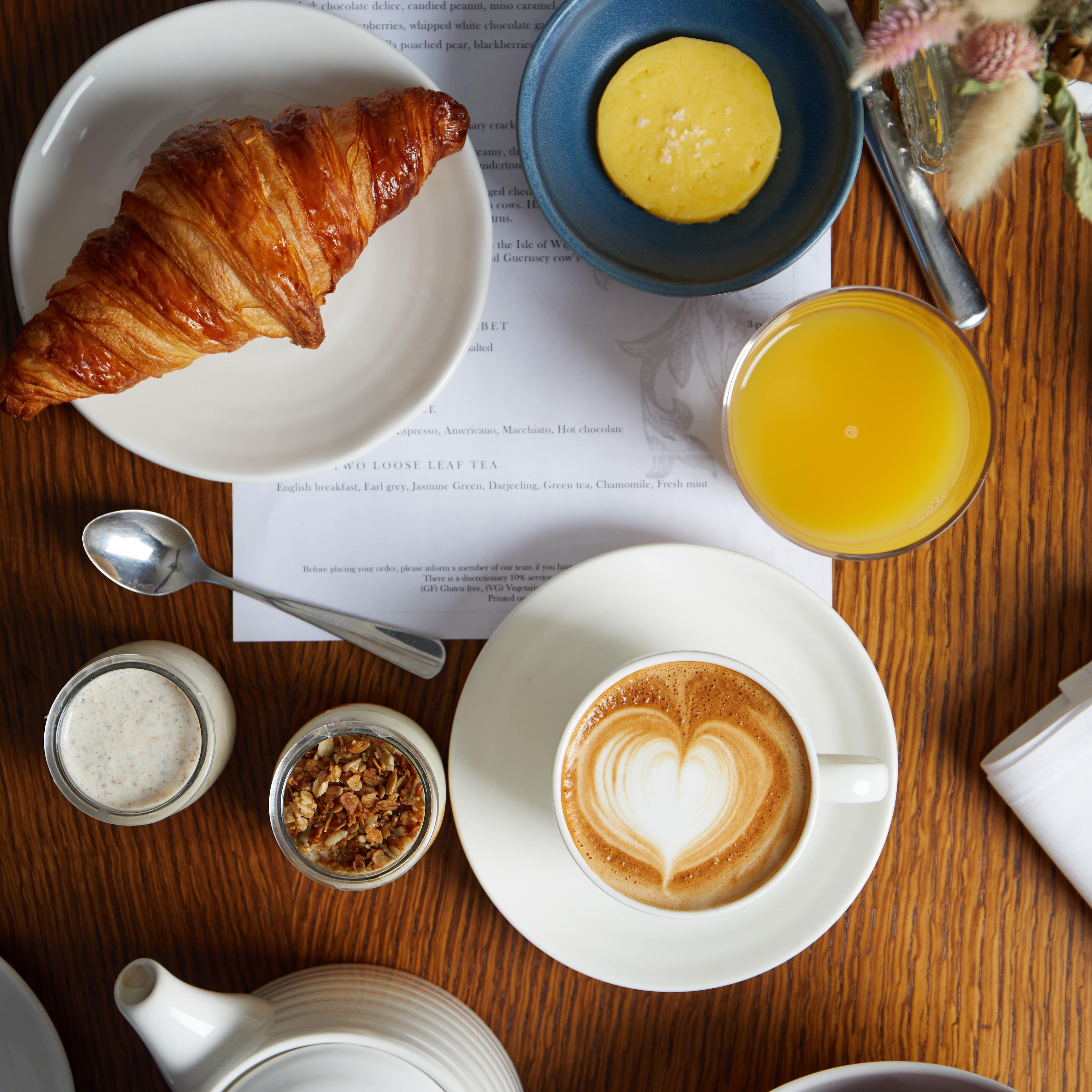 Breakfast table with croissant, butter, orange juice, cappuccino, apples, granola, and a teapot