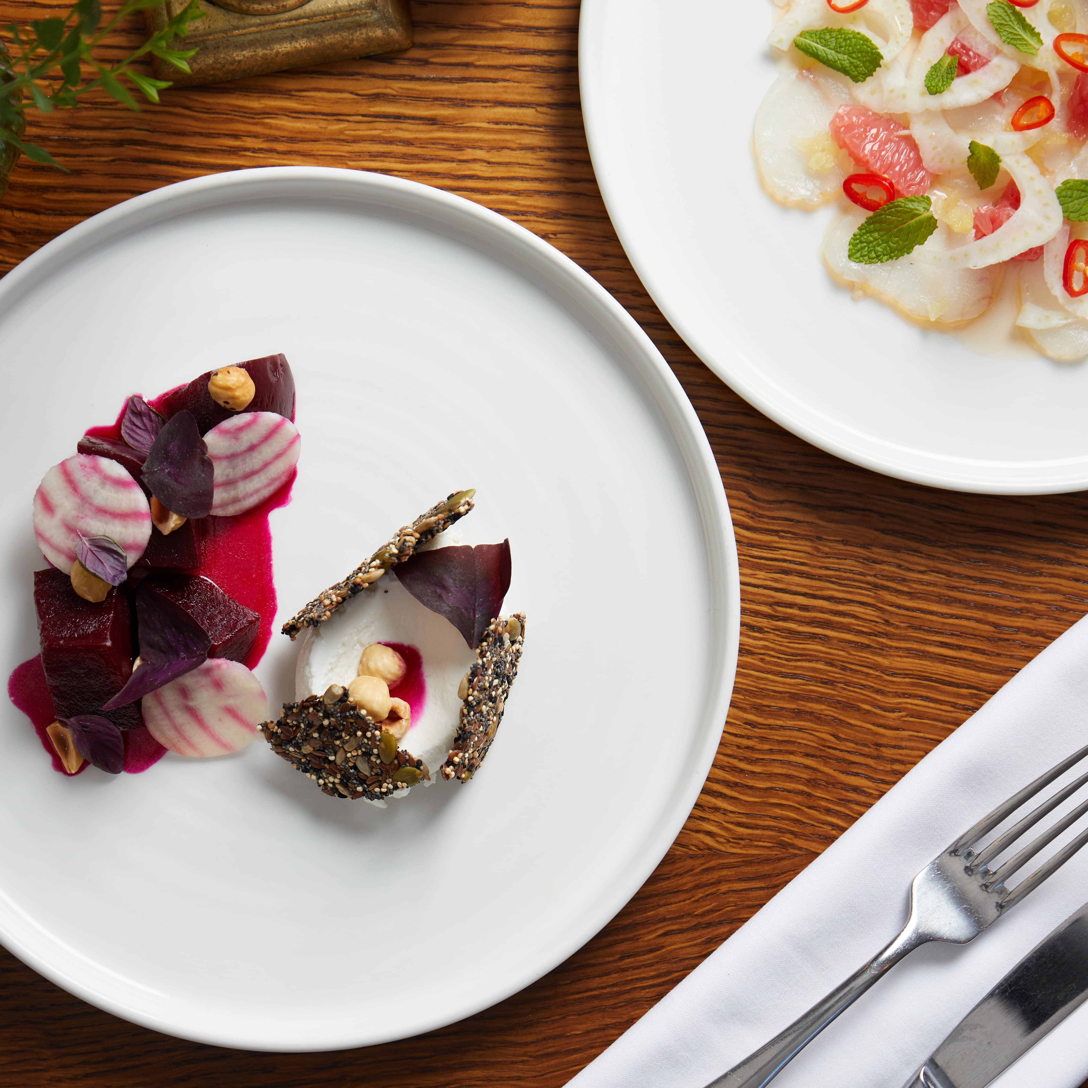 Gourmet plated dish with beets, nuts, and seeds on a white plate next to a salad on a wooden table.