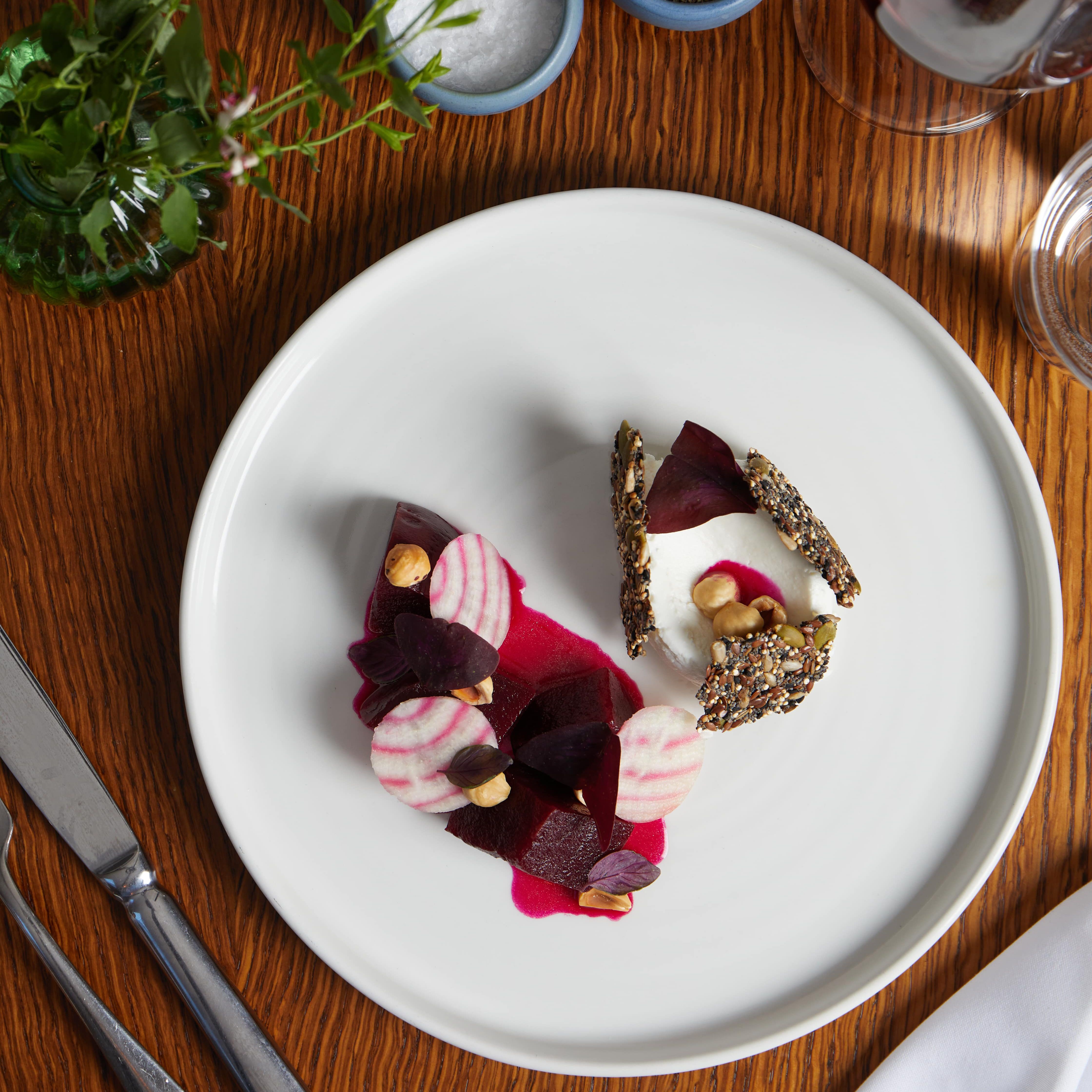A plated gourmet beet salad with nuts, goat cheese, seeds, and wafer crisps, shot from above on a wooden table with wine, water, greens, and seasoning dishes.