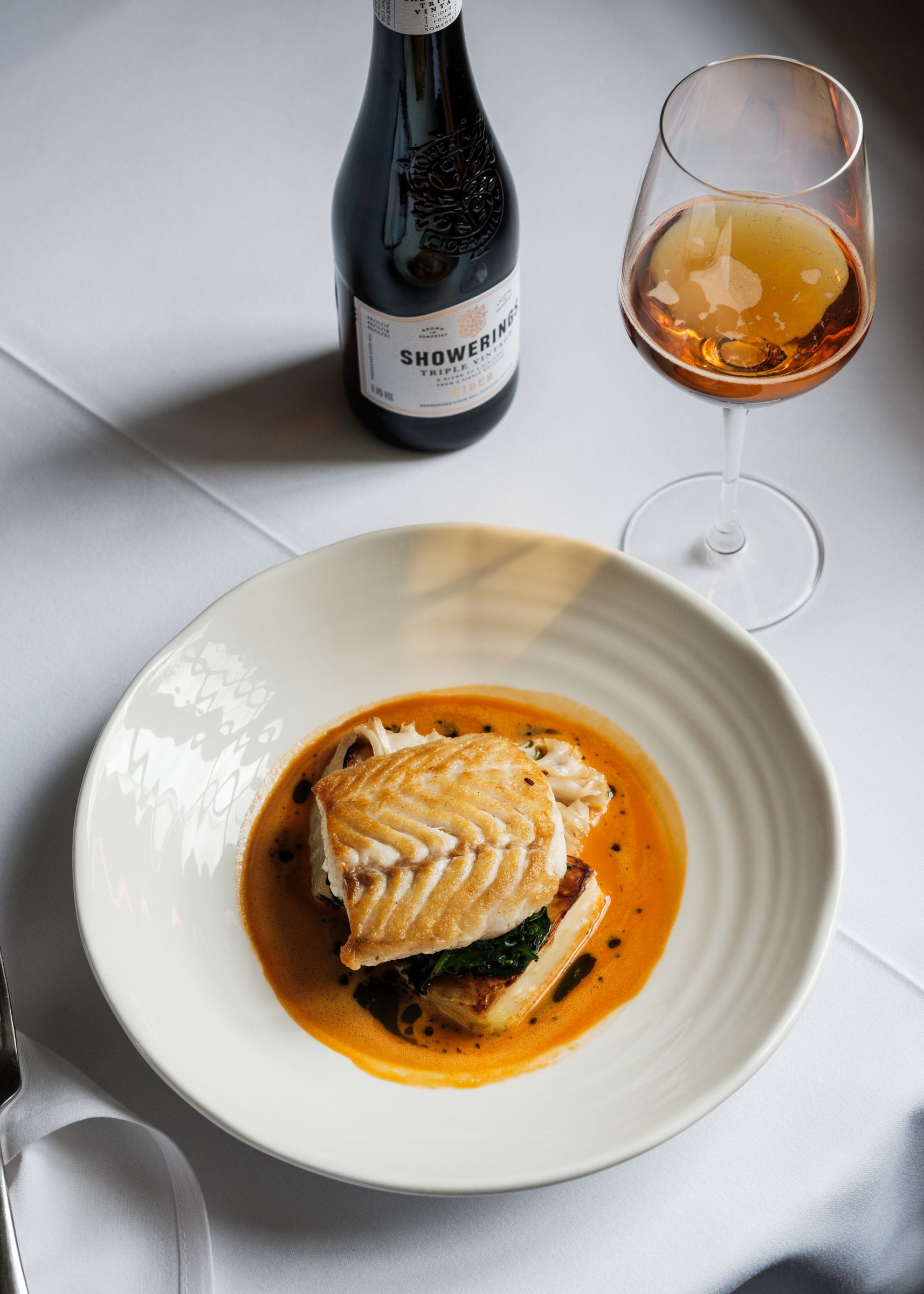 Plated fish dish with sauce, bottle of cider, and a glass of cider on a white tablecloth.