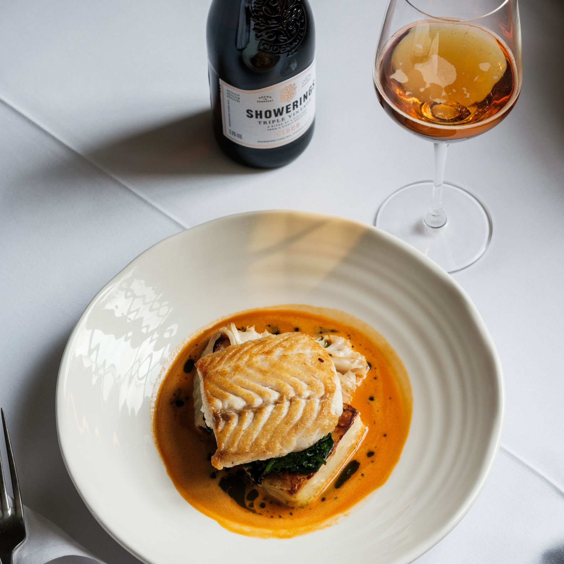 Plated fish dish with sauce, bottle of cider, and a glass of cider on a white tablecloth.