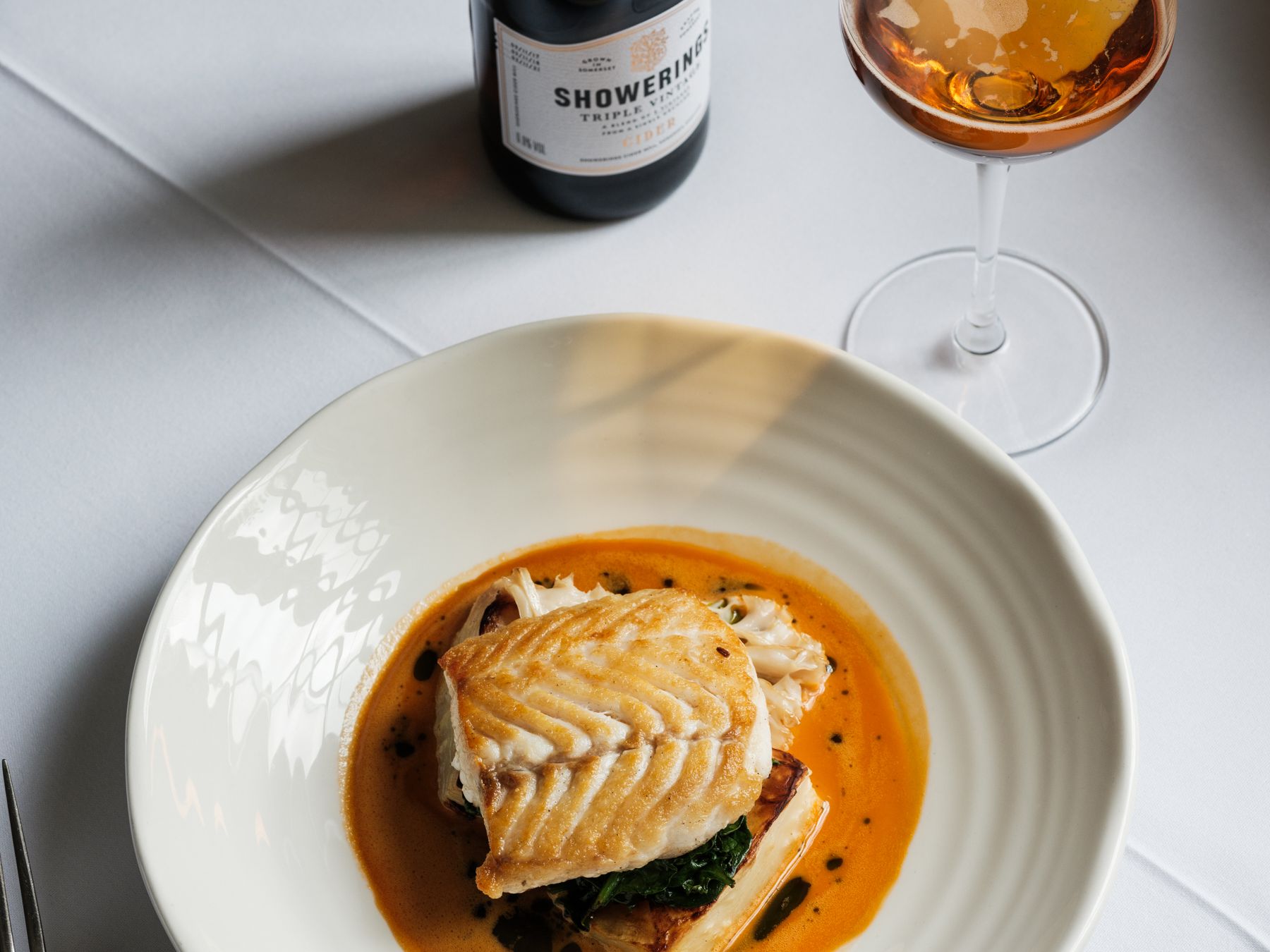 Plated fish dish with sauce, bottle of cider, and a glass of cider on a white tablecloth.
