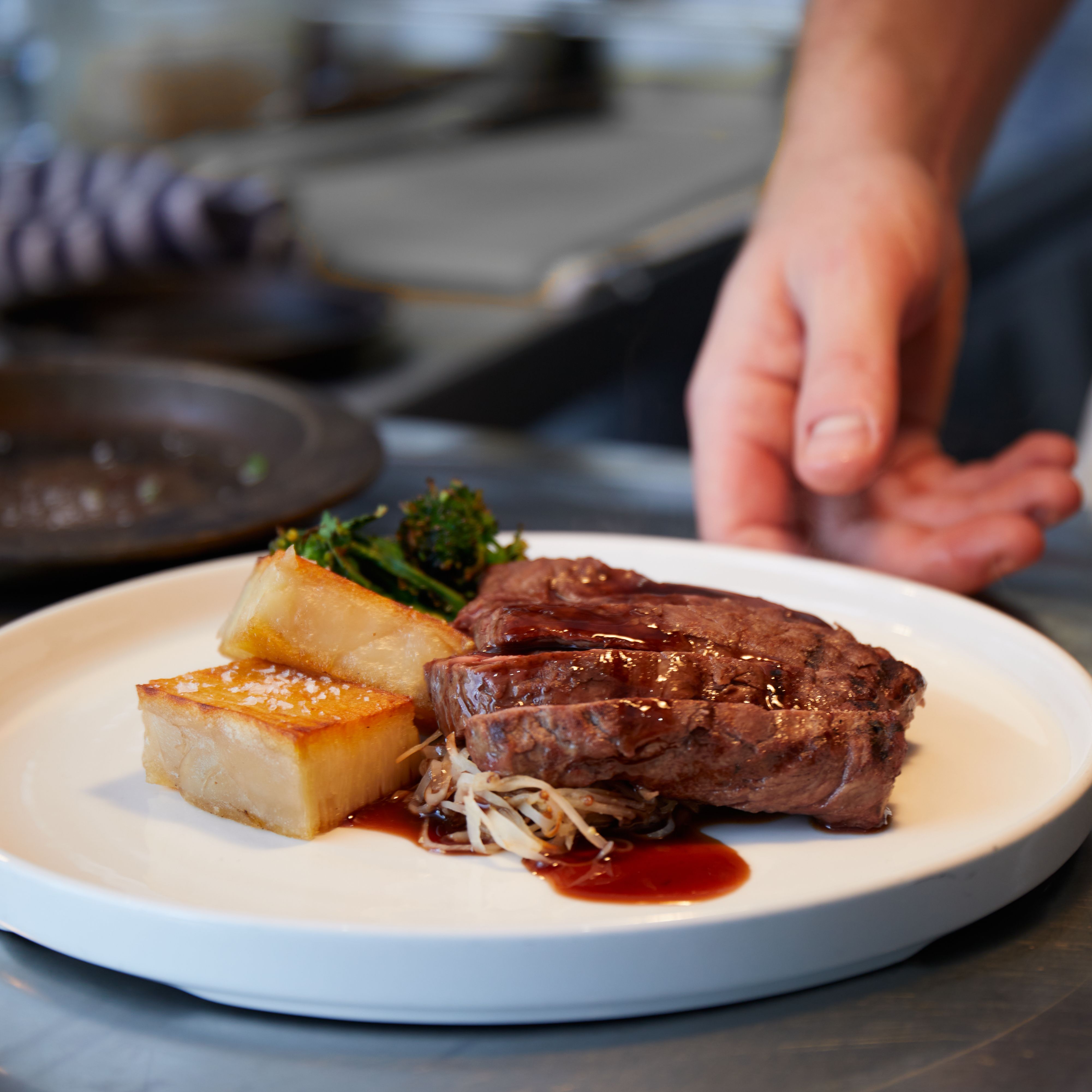 Plated steak dish with potato, greens, and sauce being prepared in a kitchen.