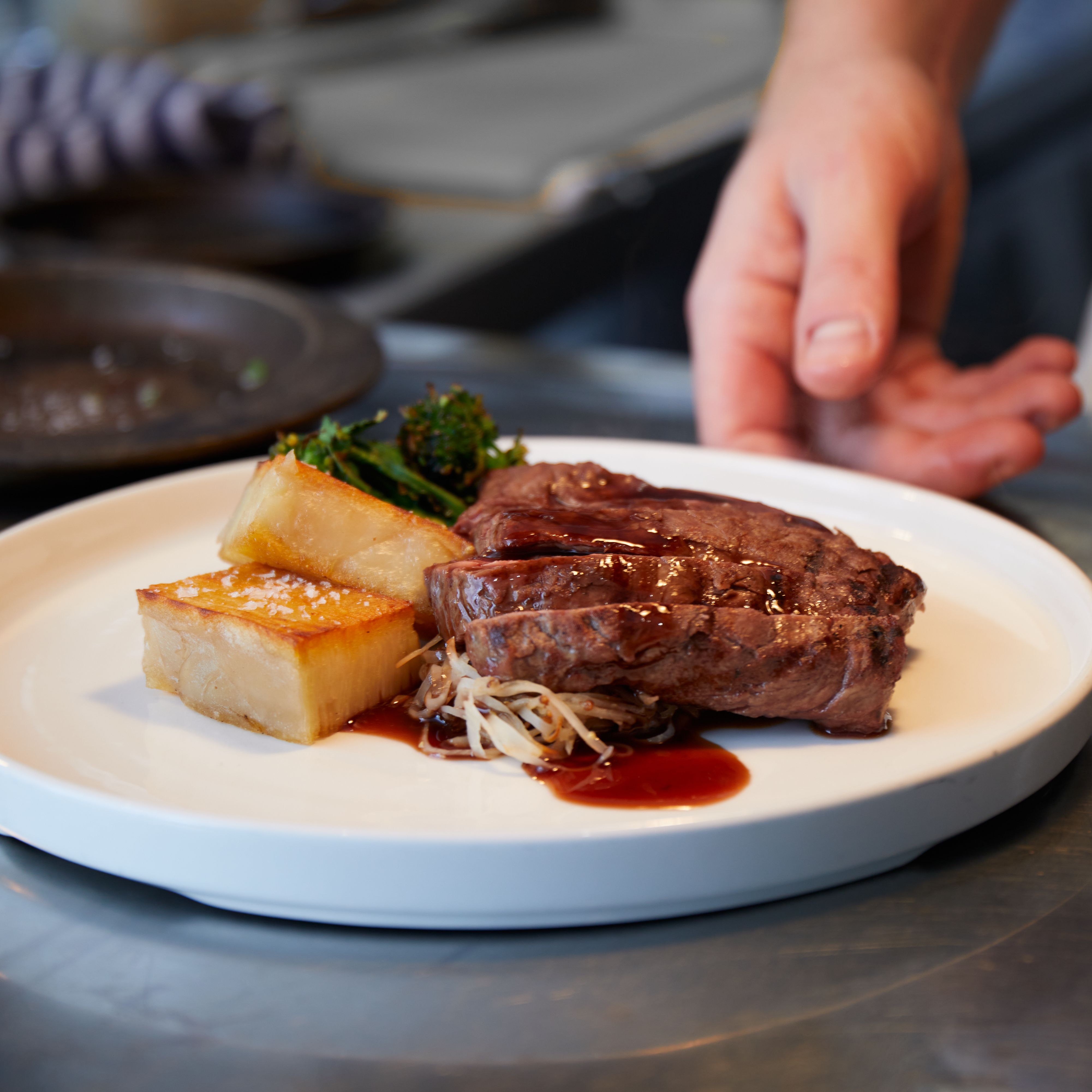 Plated steak dish with potato, greens, and sauce being prepared in a kitchen.