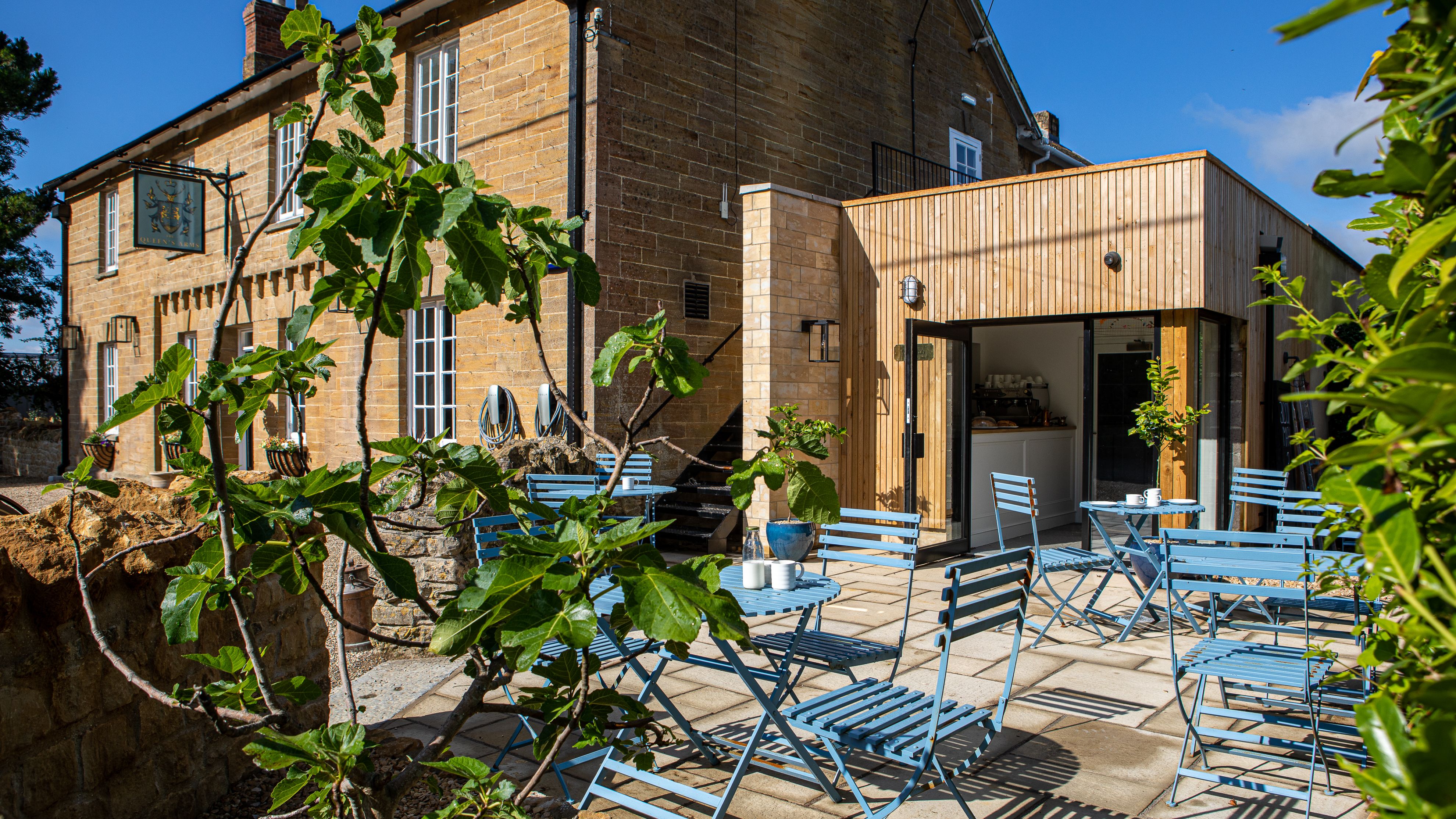 Outdoor seating area with blue chairs and tables outside a traditional stone building and modern extension.