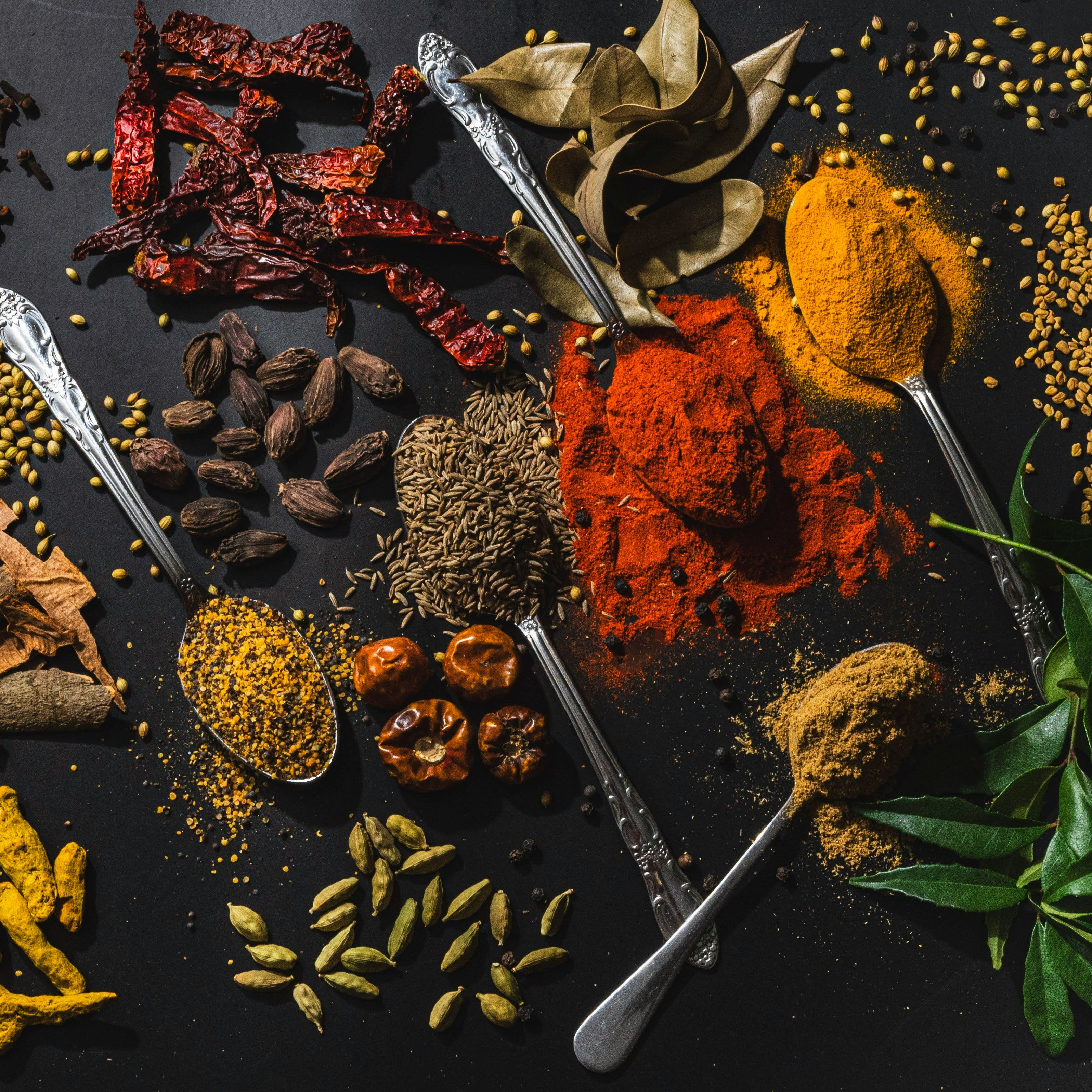 Assorted Indian spices and herbs on a dark surface with various spoons
