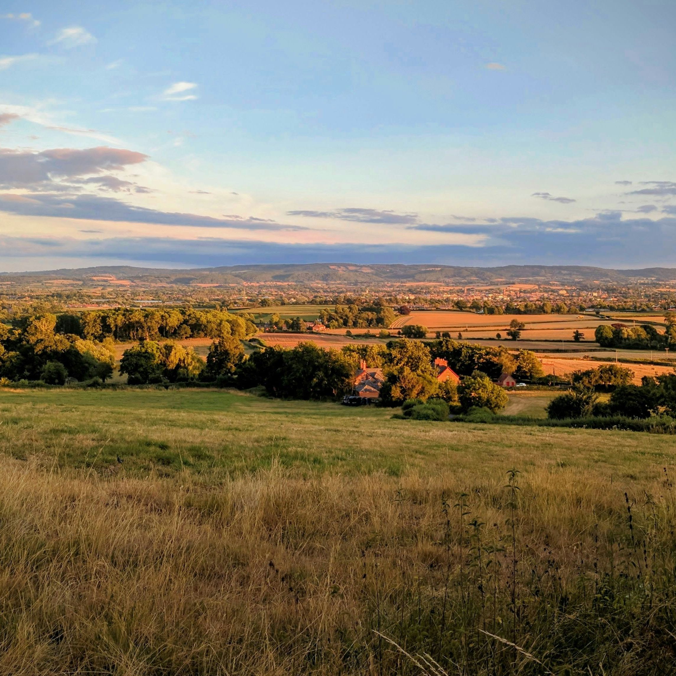 Scenic countryside landscape with rolling hills, green and golden fields, and a few houses nestled among trees under a blue sky with clouds