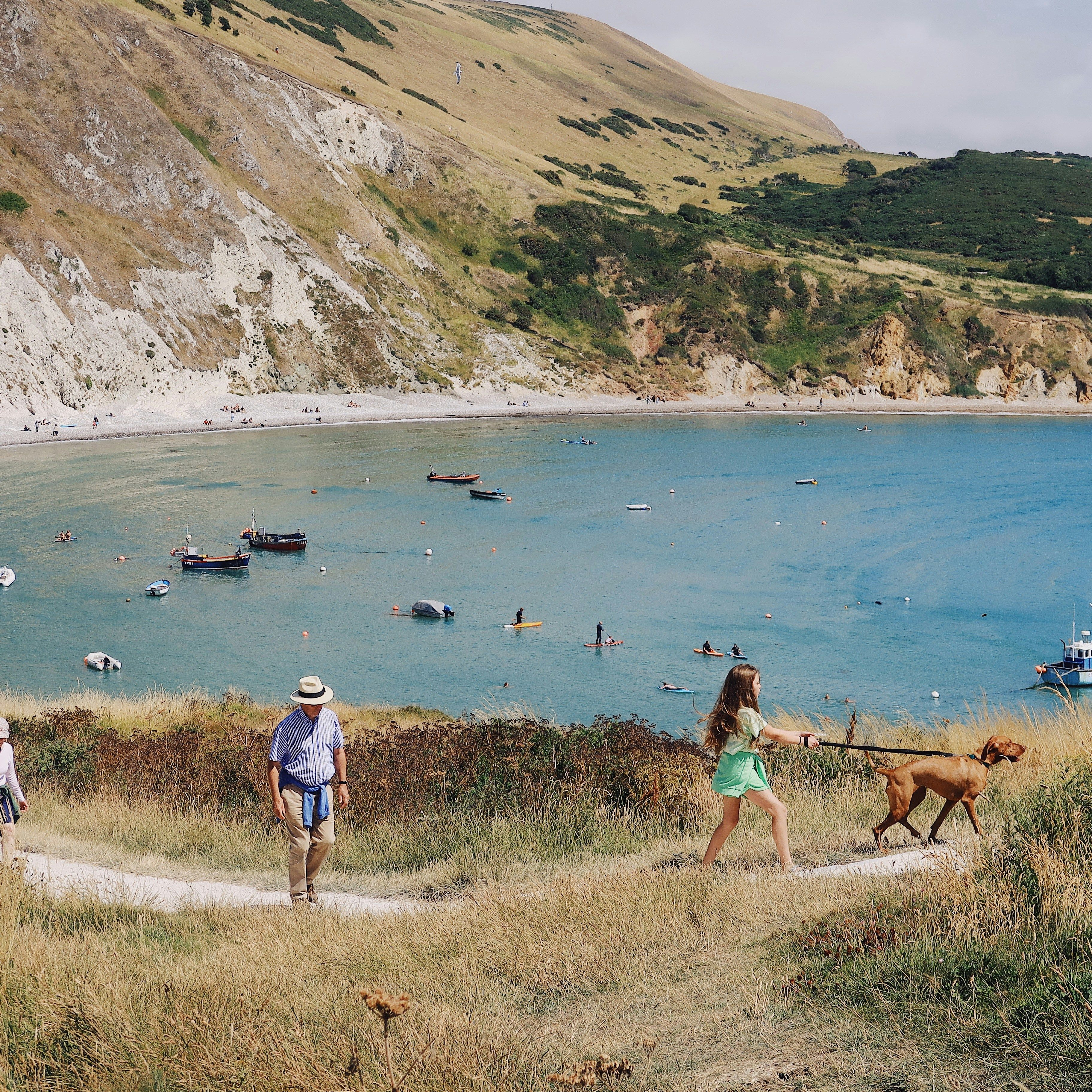 People walking along a grassy path near a turquoise bay with boats and paddle boarders, surrounded by steep rocky hills.