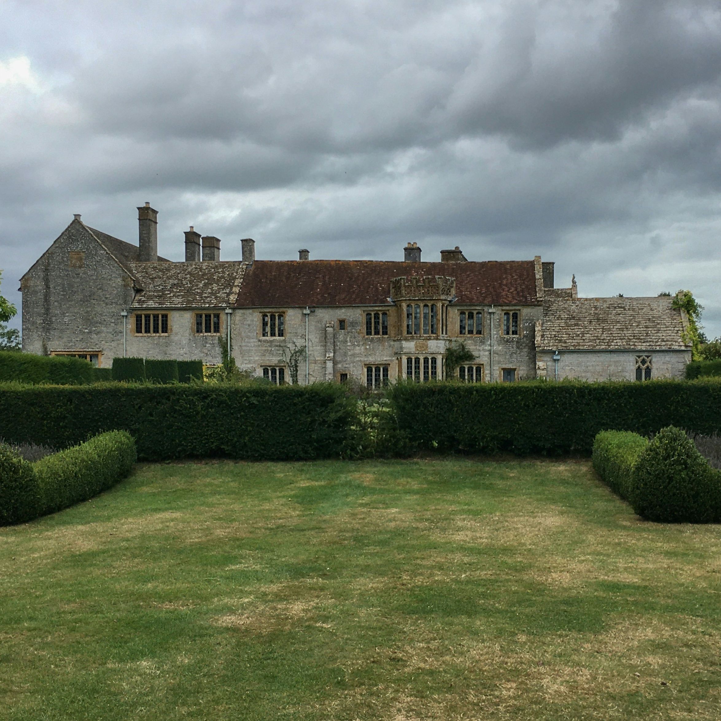 Historic stone manor house with multiple chimneys, surrounded by neatly trimmed hedges and a large lawn, under a cloudy sky.