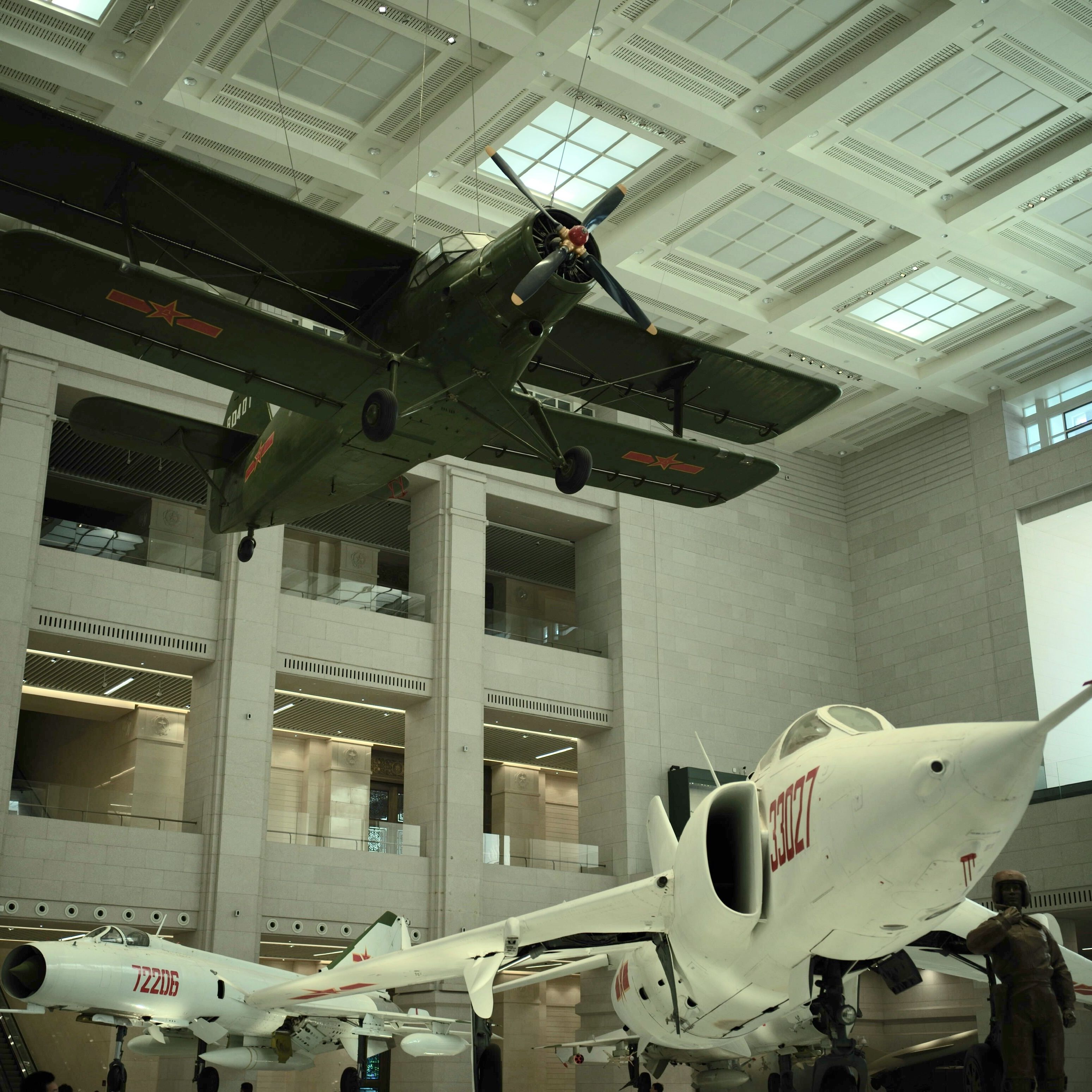 Historic military aircraft displayed in a museum with one green biplane suspended from the ceiling and a white jet fighter beneath it.