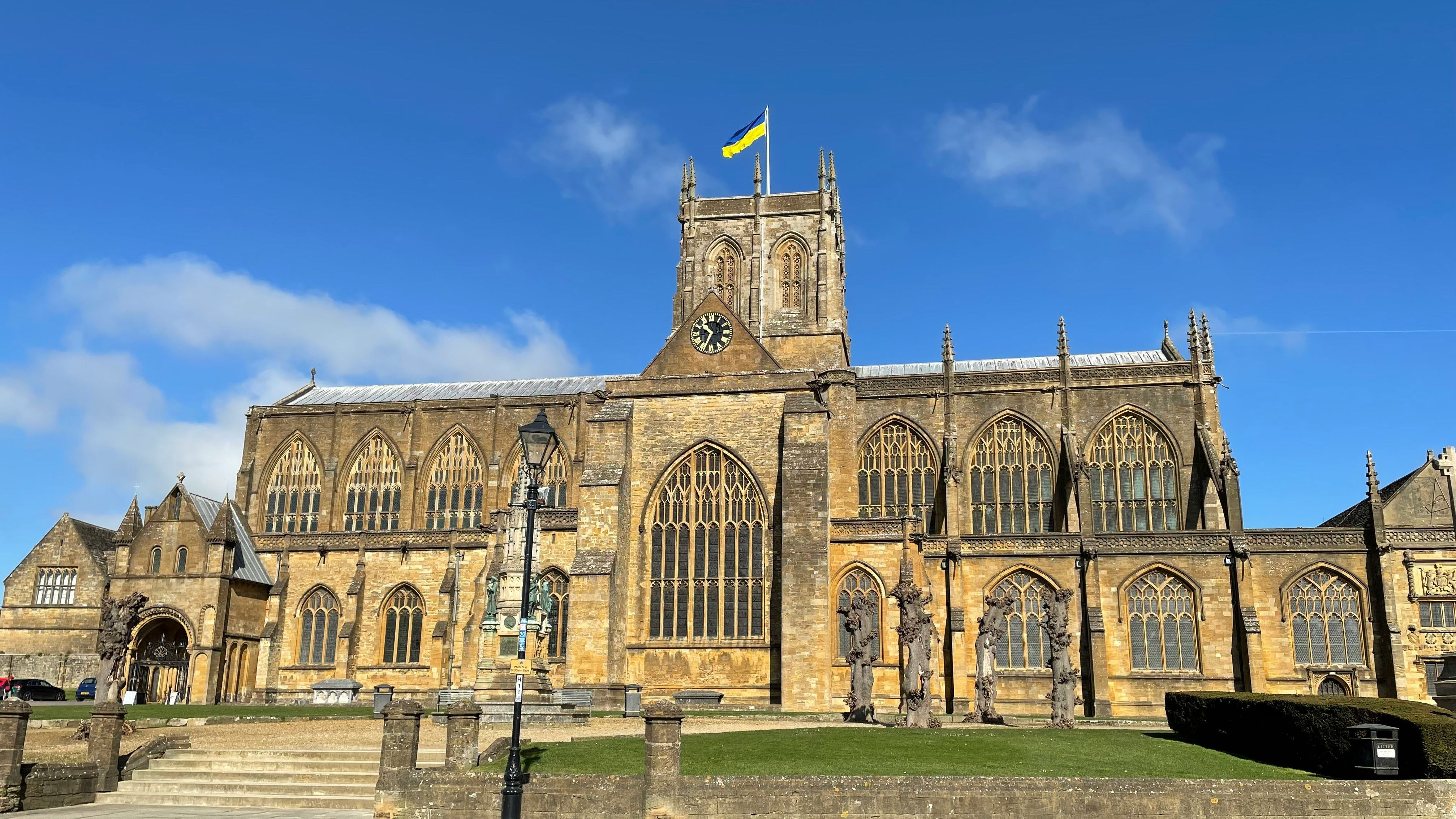 Historic stone church building with arched windows, a clock tower, and a blue and yellow flag on top, under a blue sky.