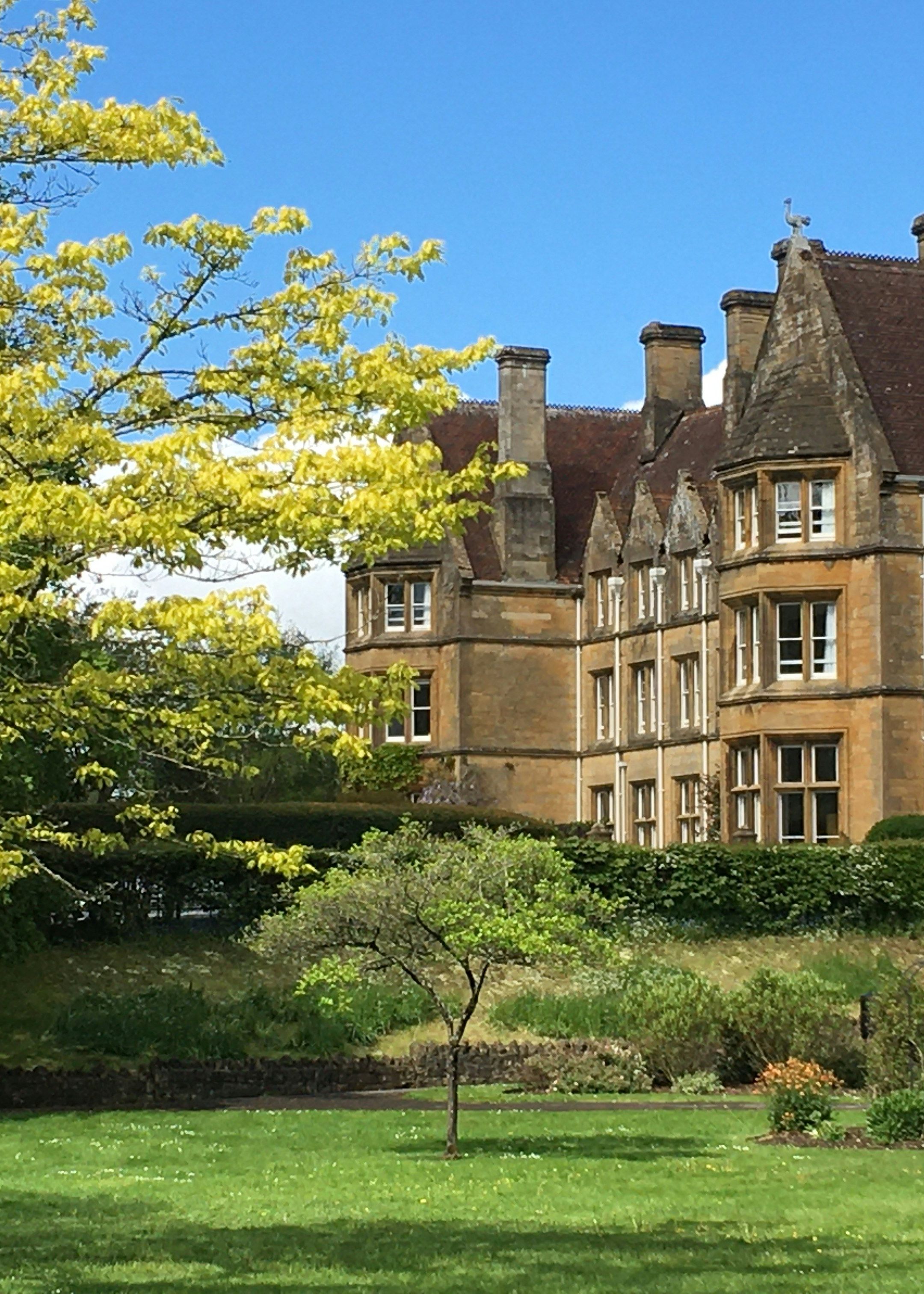 Historic stone building with multiple chimneys and steep roofs, set behind green lawns and trees on a sunny day.