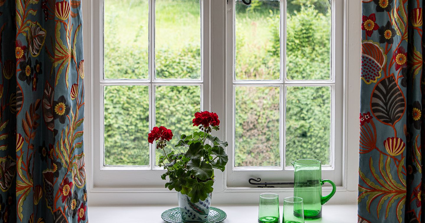 A windowsill with a potted red geranium and green glassware in front of a garden view.