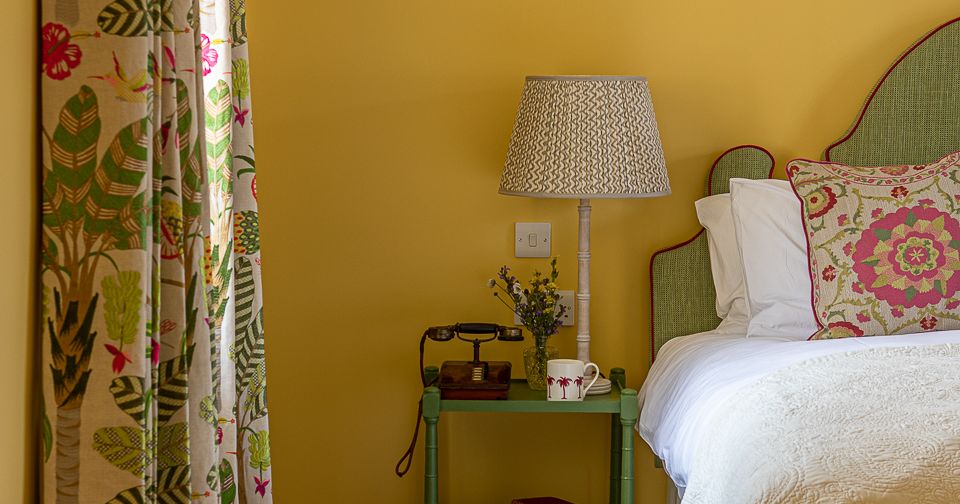 Cozy bedroom corner with floral curtains, a green nightstand, bedside lamp, old-fashioned telephone, books, and a bed with a patterned cushion.