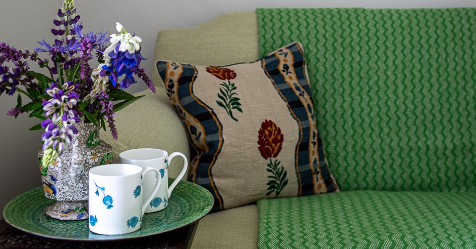 Cozy living room corner with green sofa, decorative pillow, flower vase, two mugs, and framed landscape painting.