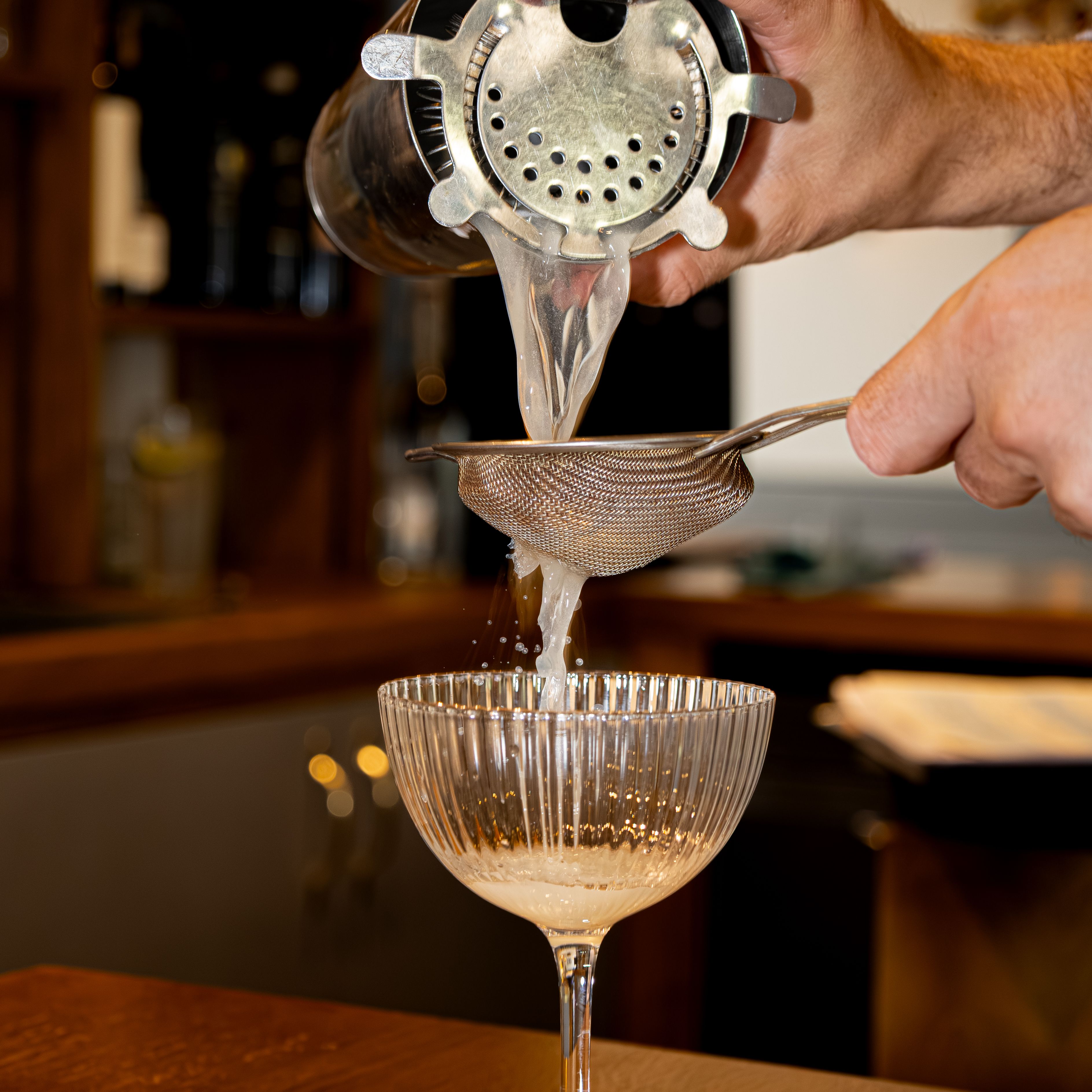 Bartender pouring a cocktail through a strainer into a coupe glass