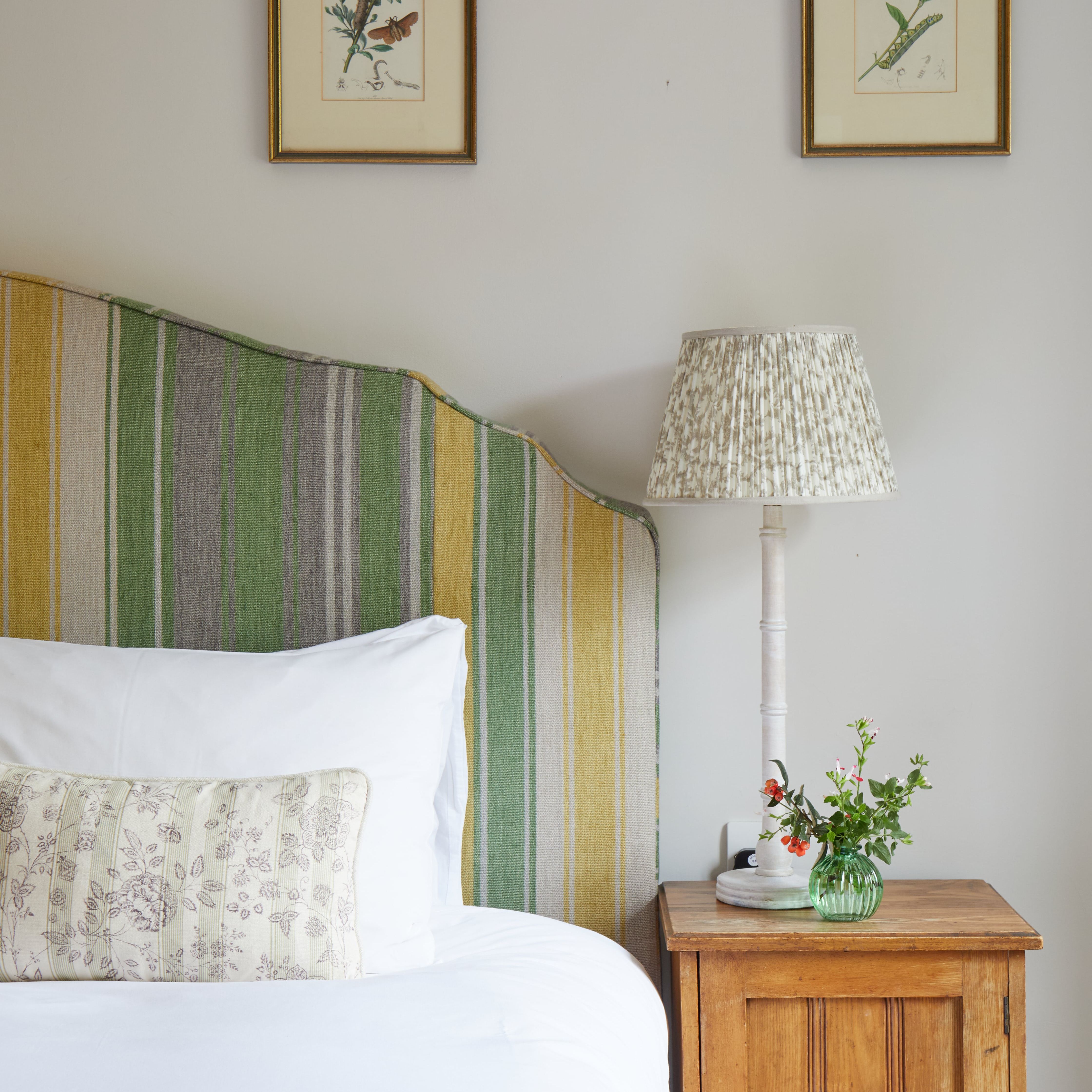 A cozy bedroom corner with a striped headboard, white bedding, wooden nightstand, table lamp, and two botanical prints on the wall.