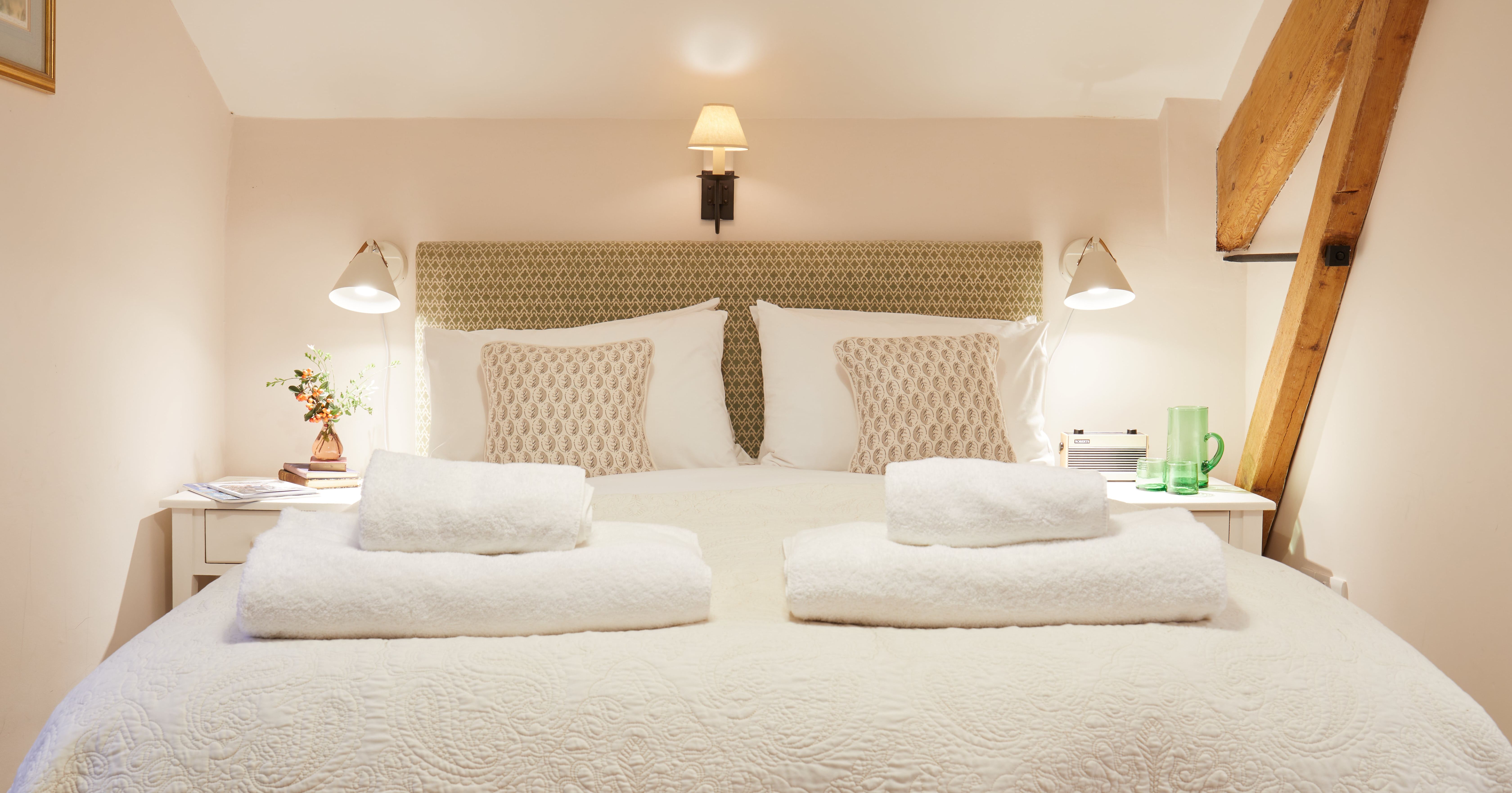 Cozy and inviting bedroom with a double bed decked in white linens and folded towels, flanked by bedside tables, lamps, vintage decor, and rustic wooden beam.
