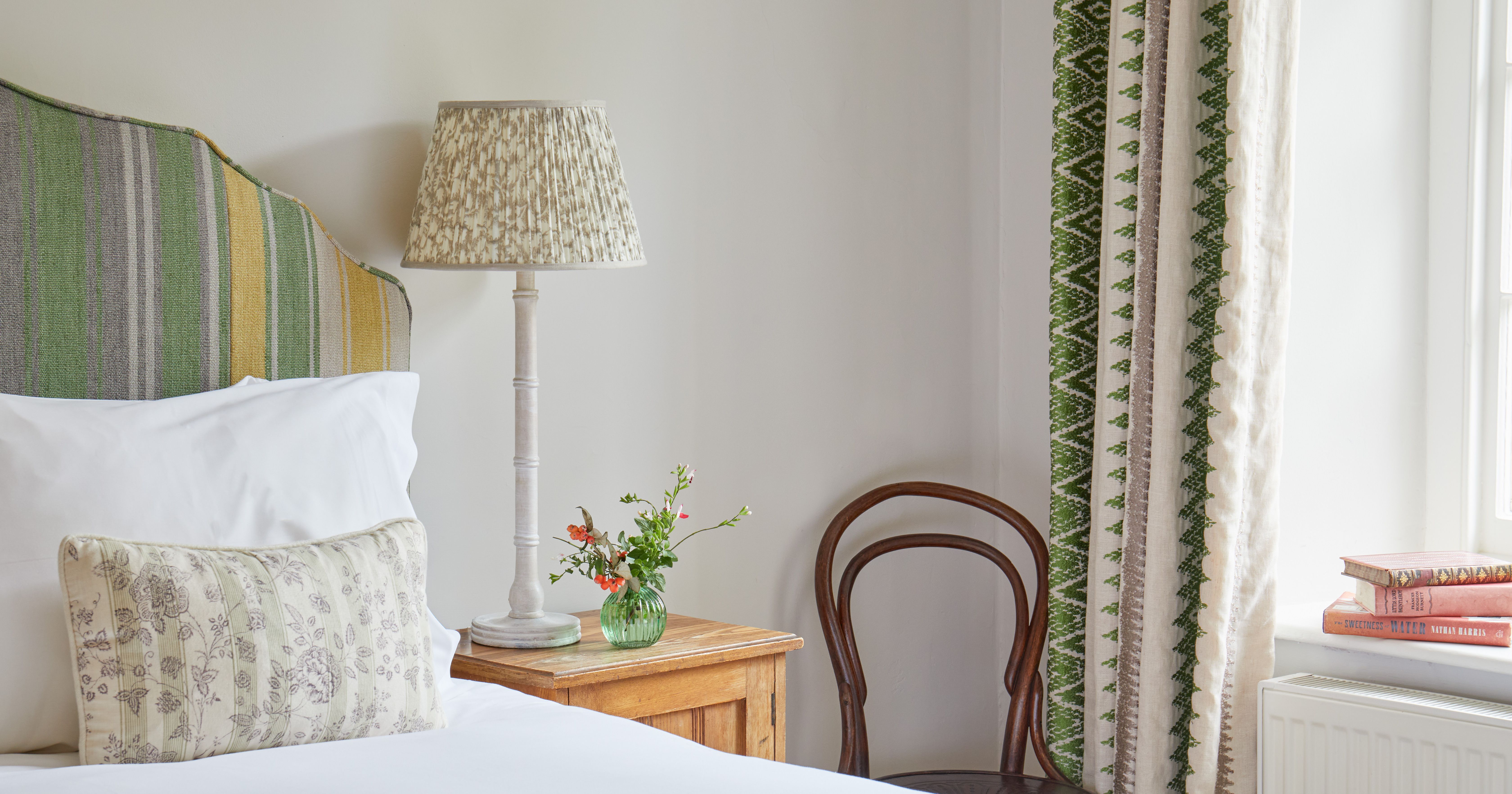 Cozy bedroom corner with striped headboard, bedside table, lamp, chair, flowers, and books.