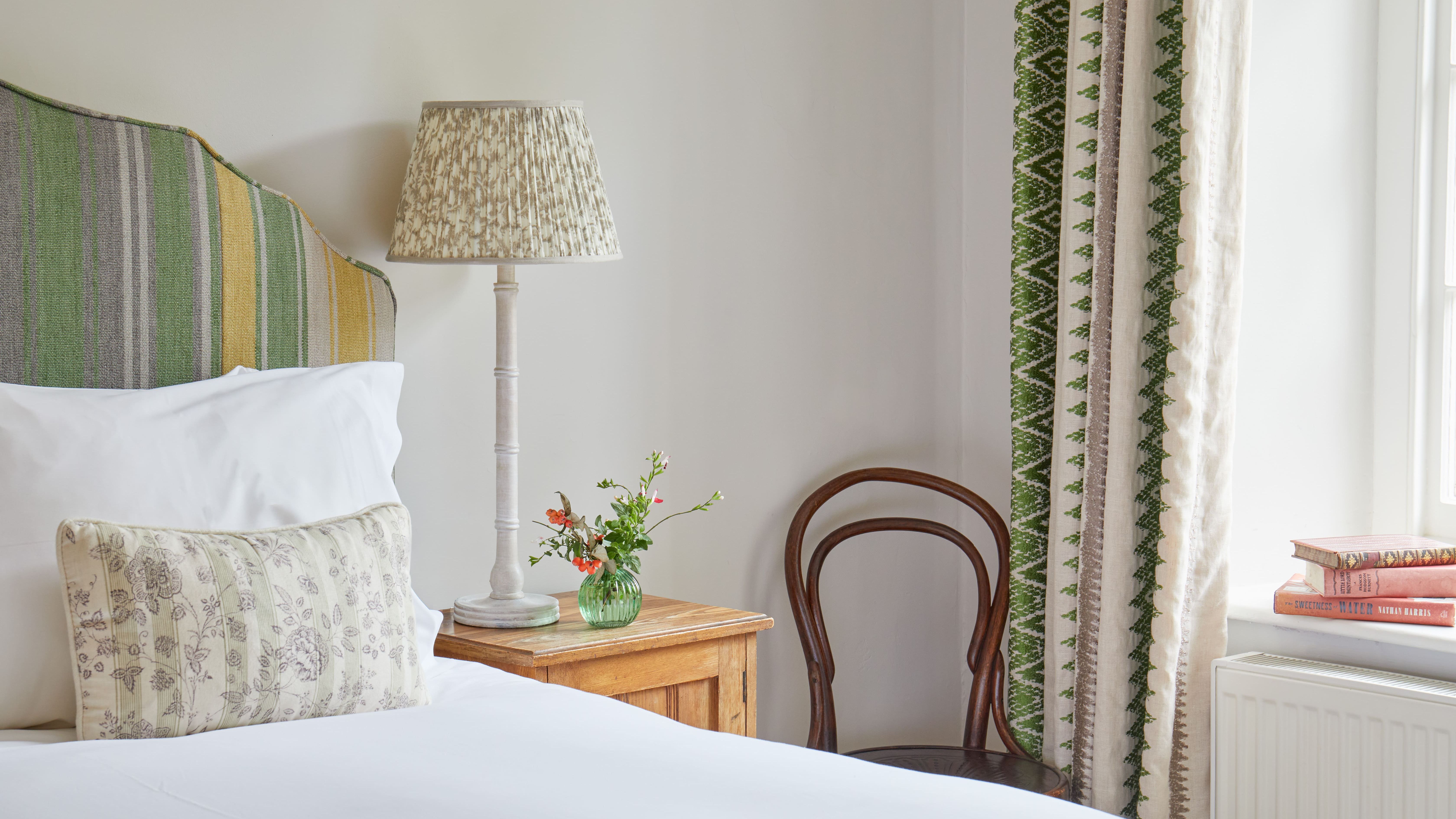 Cozy bedroom interior with a striped headboard, bedside lamp, small vase with flowers, wooden chair, and patterned curtains