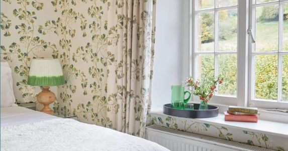 Cozy bedroom with floral wallpaper, white bedding, and a window seat with a tray, books, and flowers.