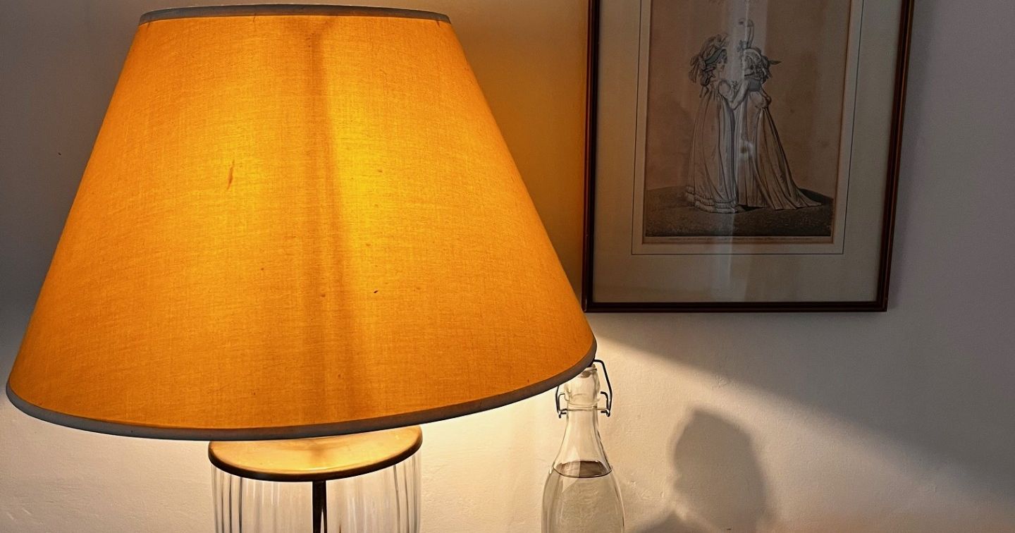 A warmly lit table with a lamp, two glasses, a glass bottle, and a basket of tea and snacks.