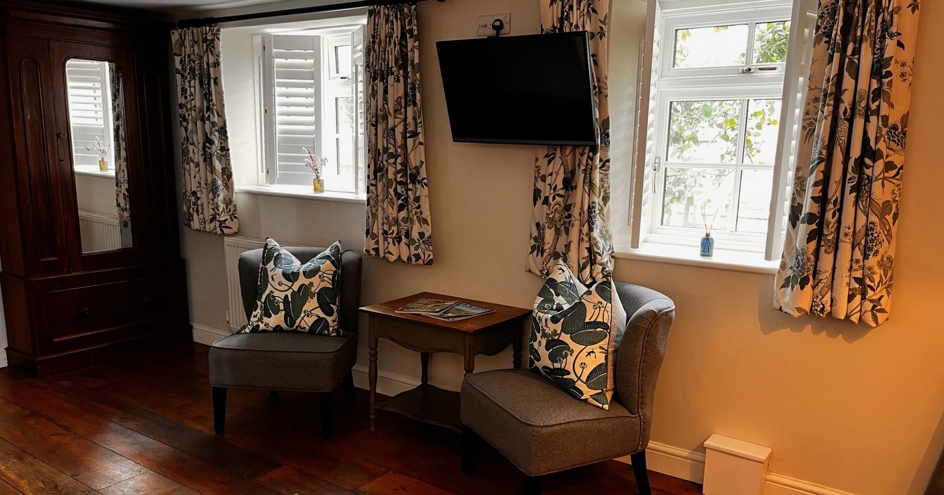 Cozy corner of a living room with two gray chairs, a small wooden table, floral curtains, and a TV mounted on the wall.
