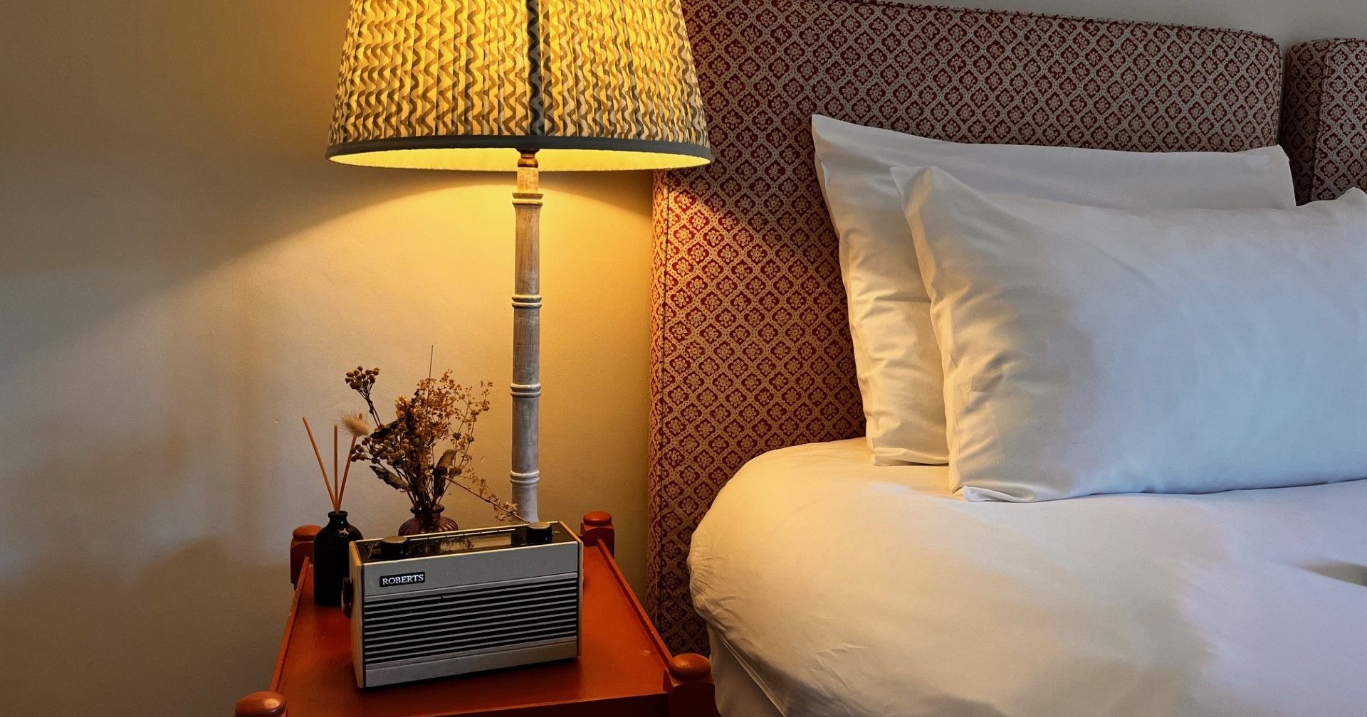 A cozy bedroom scene with a lit table lamp, a bedside table with decorative items, and a neatly made bed with white pillows.