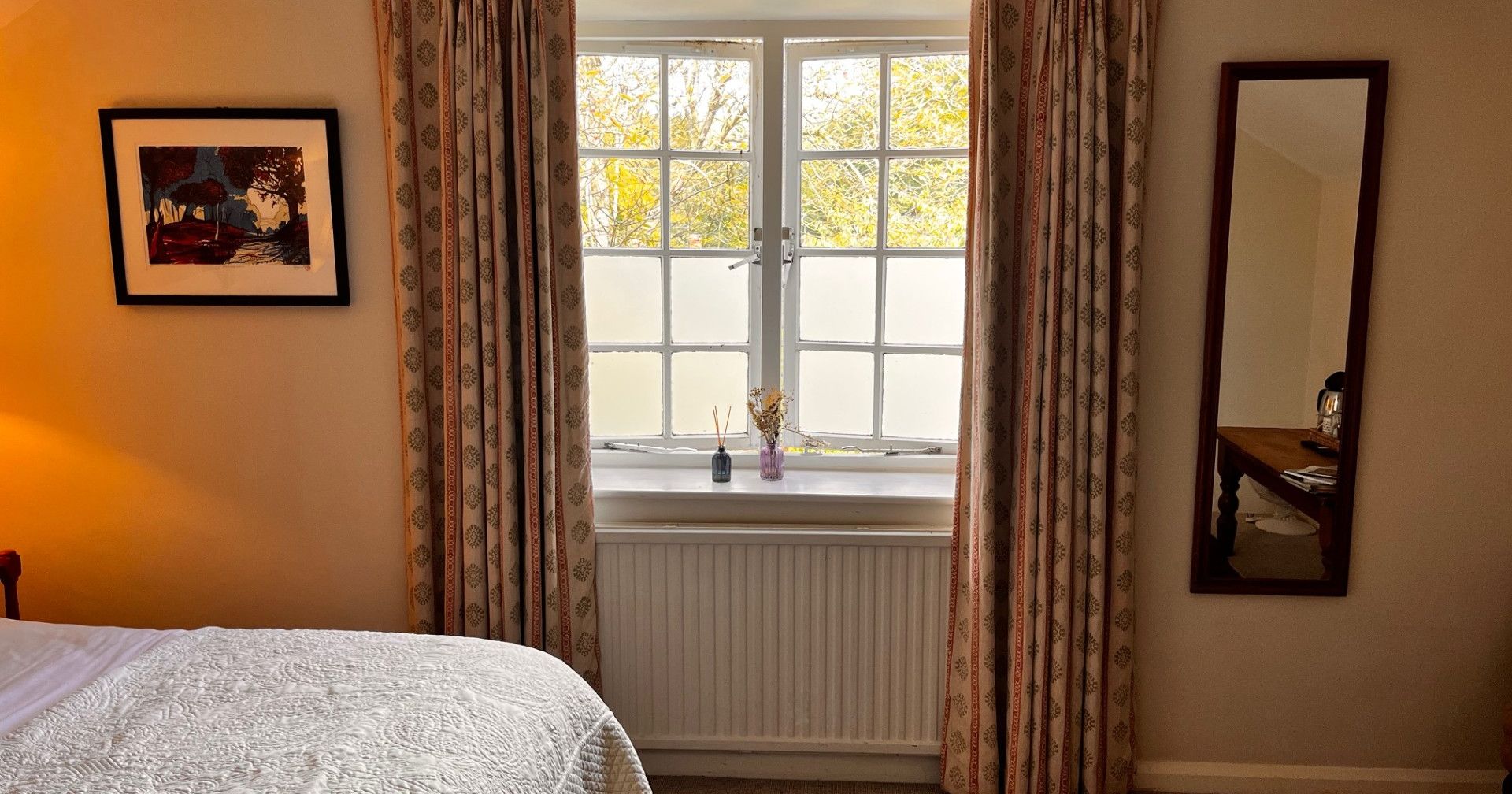 Bedroom window with floral curtains, a bed, framed art, and a large wall mirror.