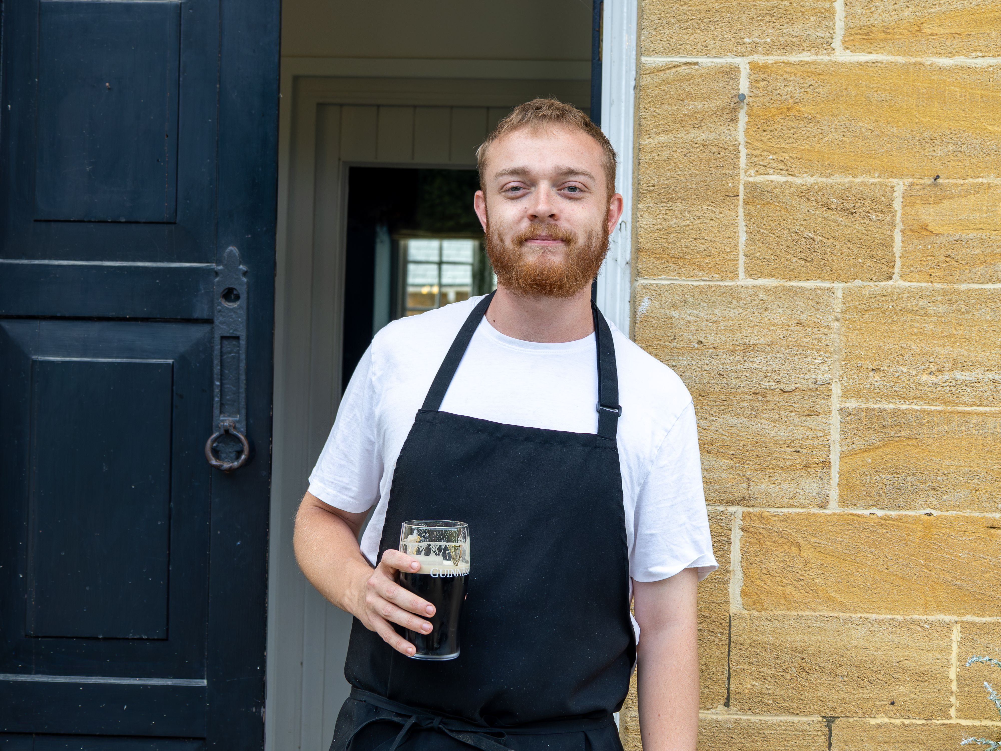 Man in a black apron holding a pint of dark beer standing by a doorway.