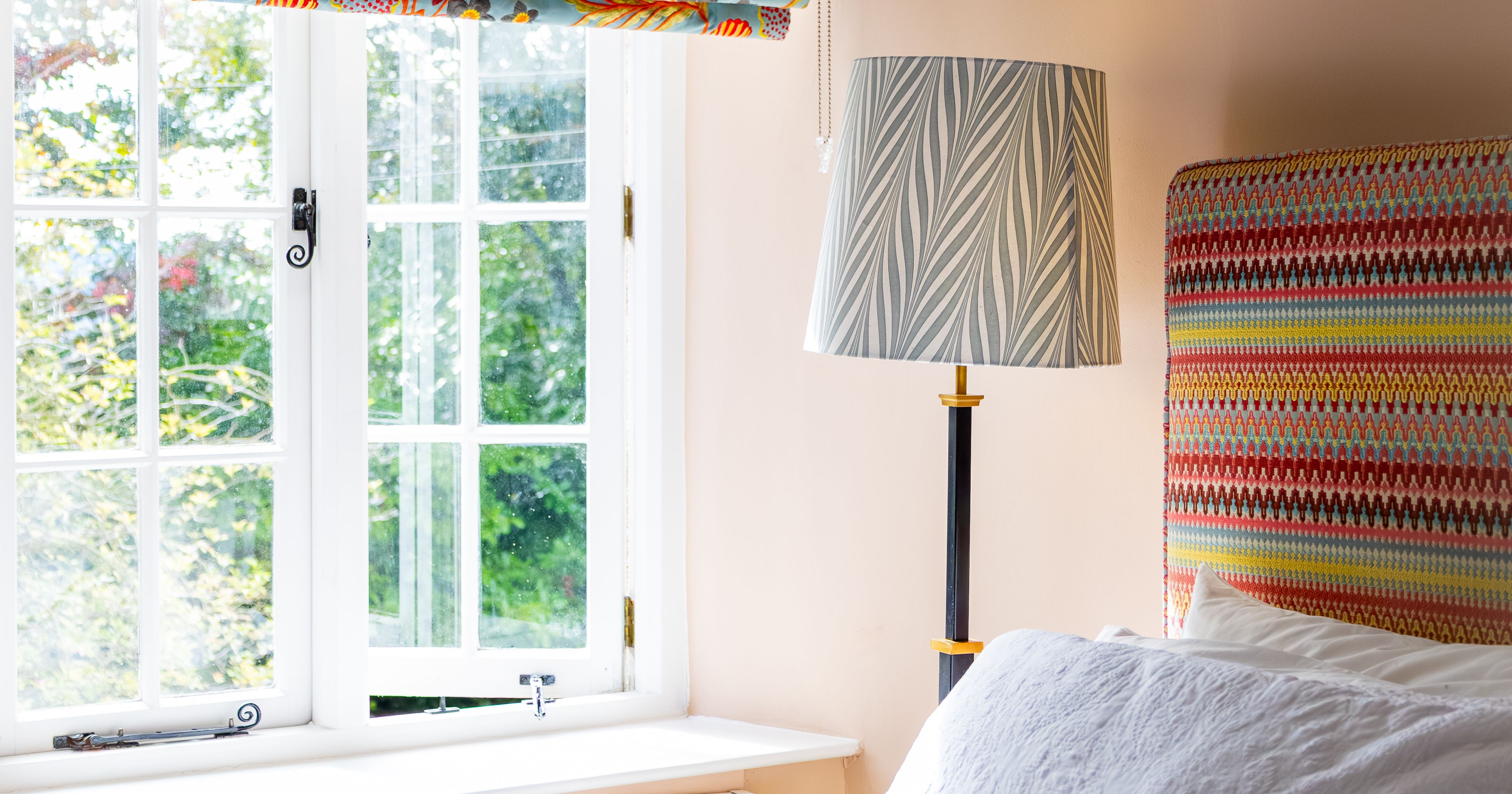 Brightly lit bedroom with a colorful patterned window shade, a bed with white linens, a lamp with a leaf-patterned shade, and a patterned headboard.