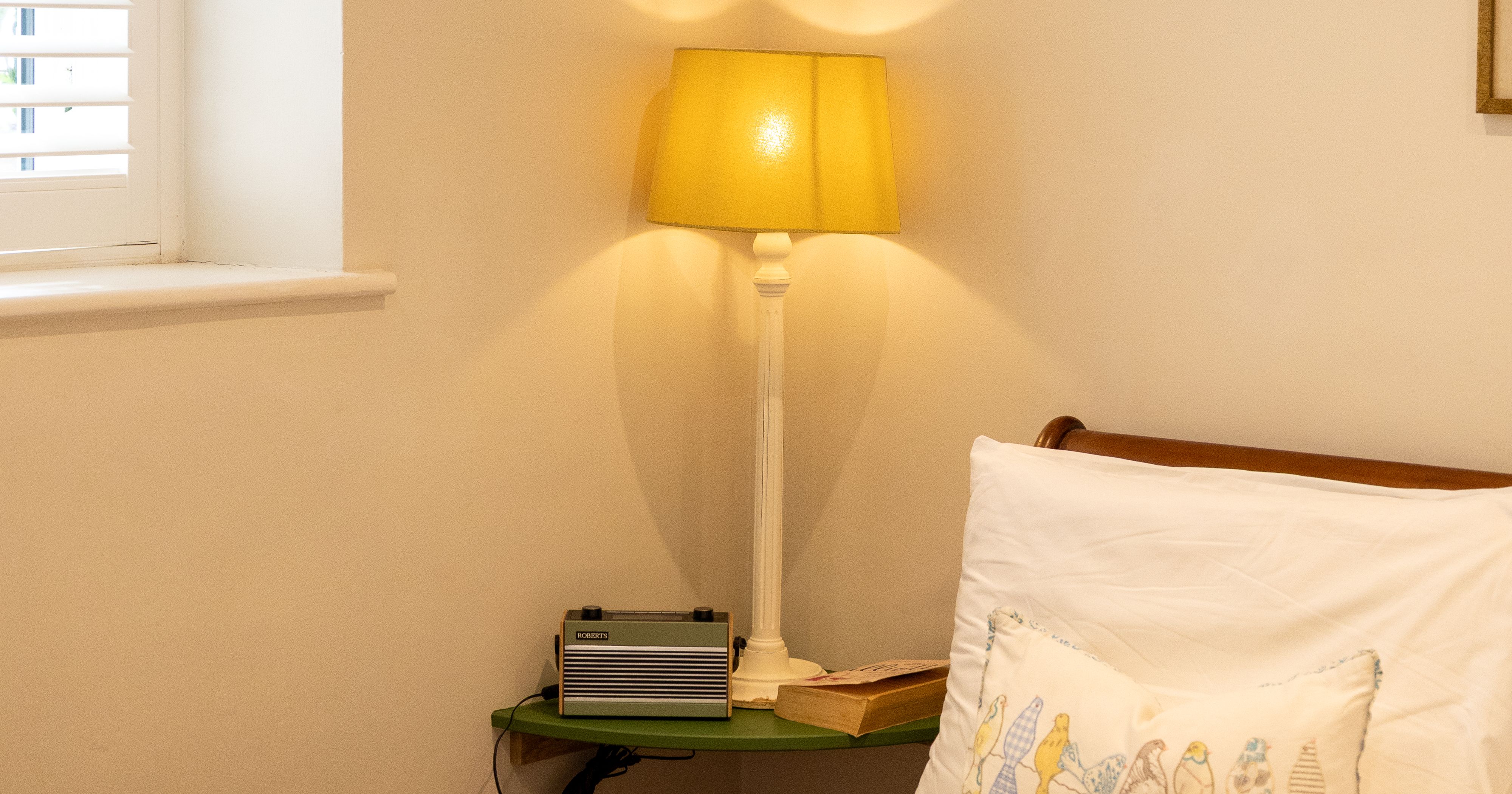 A cozy bedroom corner with a yellow lamp, radio, and a pillow featuring birds on a white bed.