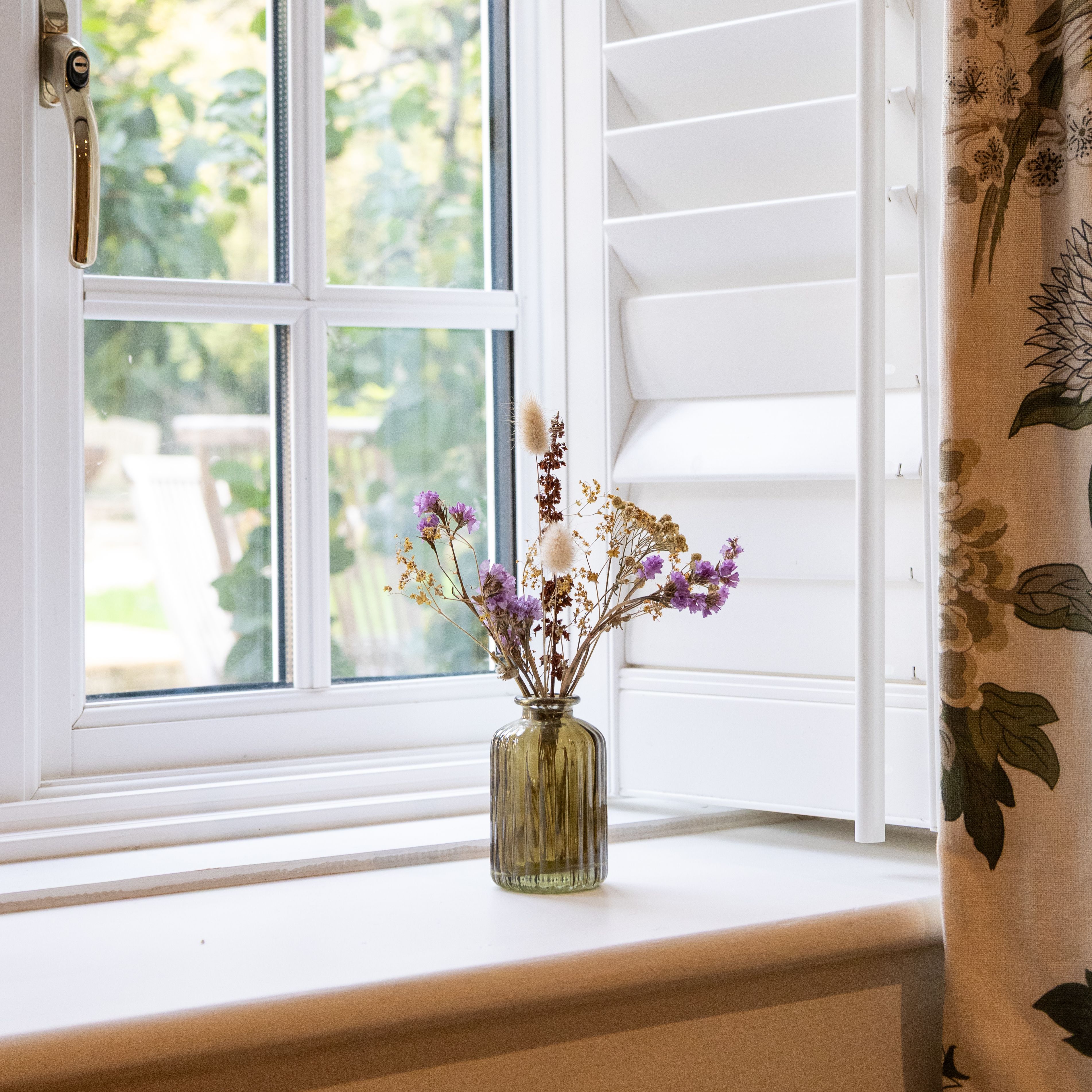 A small vase with dried flowers sitting on a windowsill beside a window with white shutters.