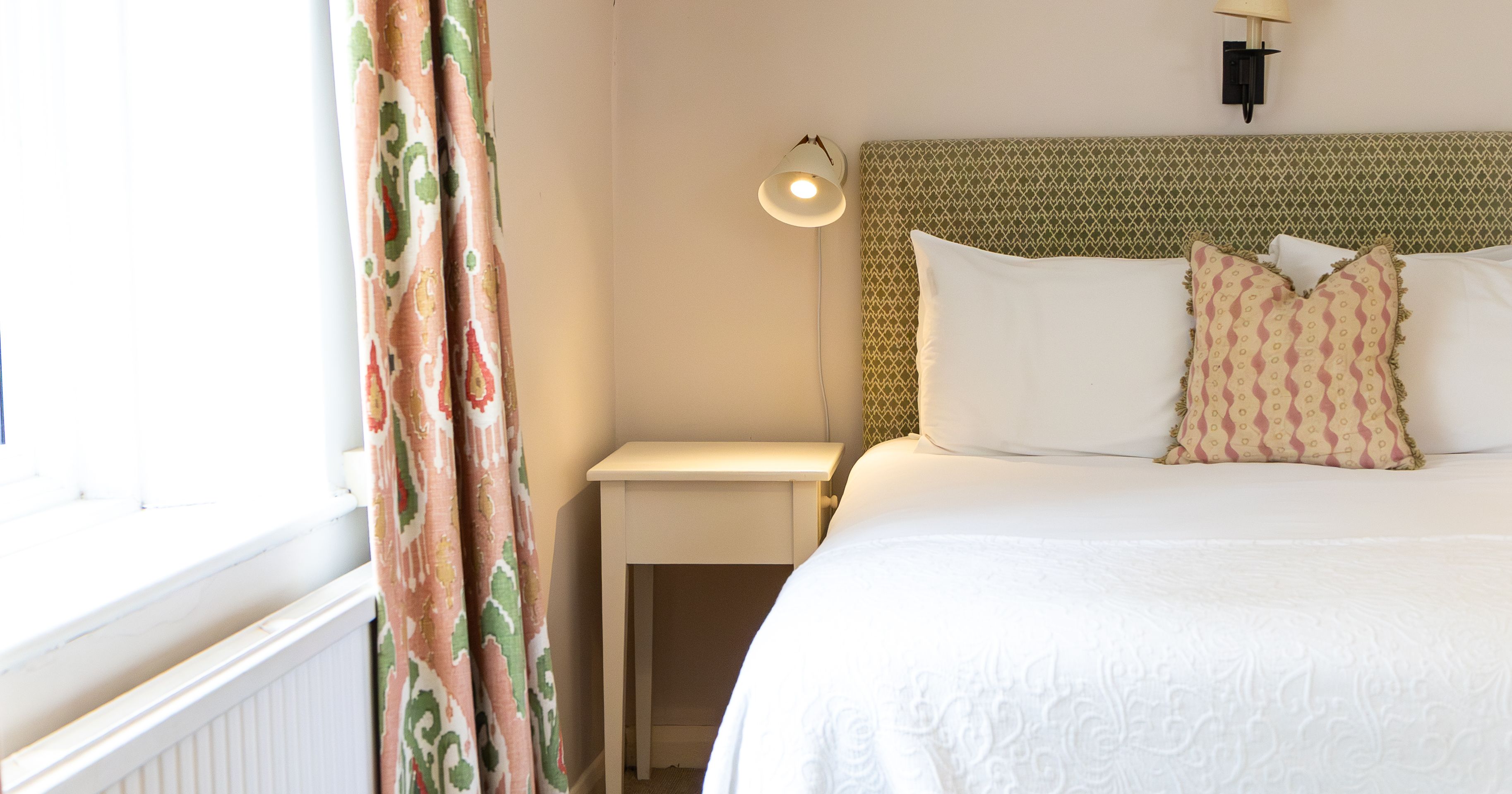 Bright bedroom with a neatly made bed, patterned curtains, a bedside table with a lamp, and a window letting in natural light.