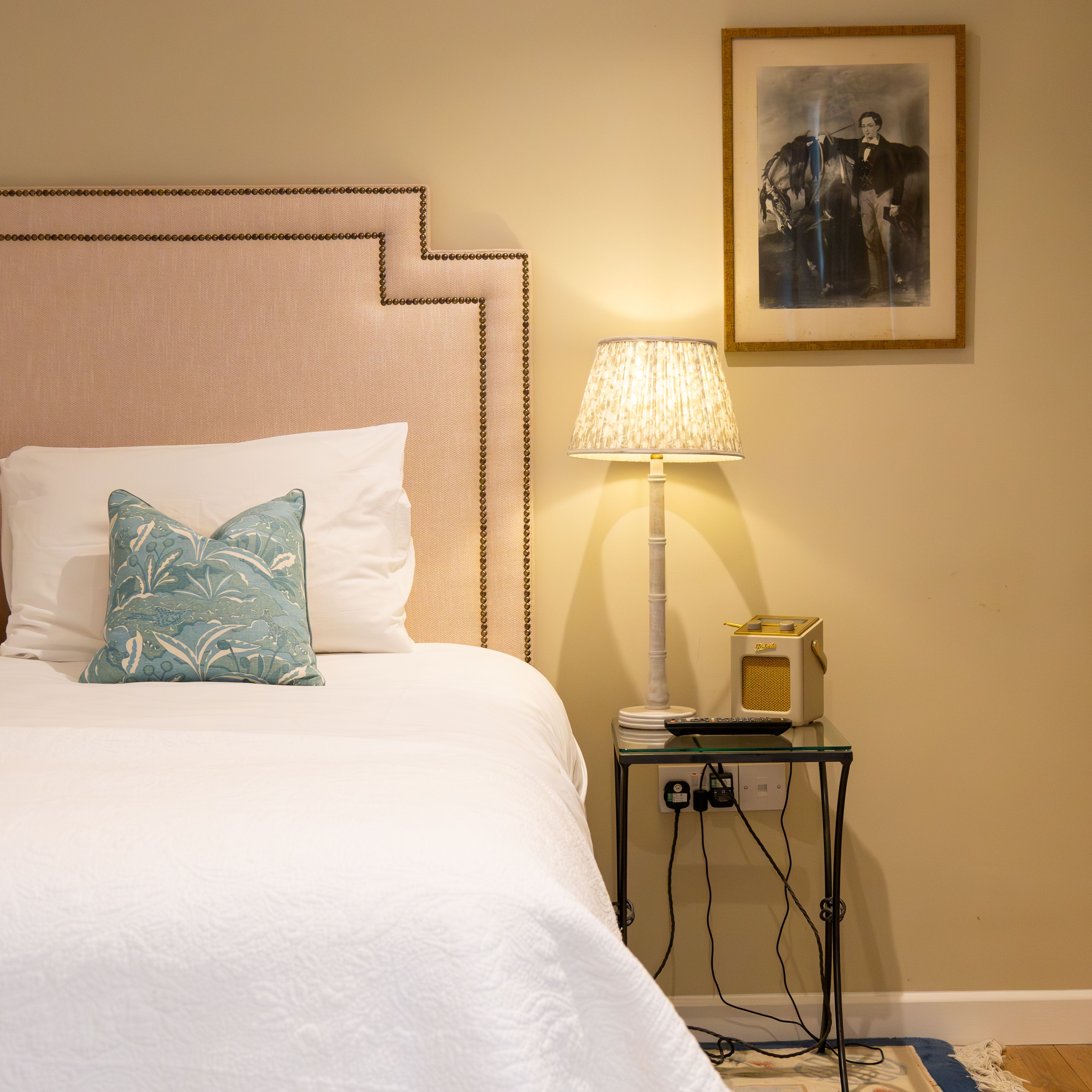 Cozy bedroom with a large bed, two teal pillows, a nightstand with a lamp, and framed artwork on the wall.