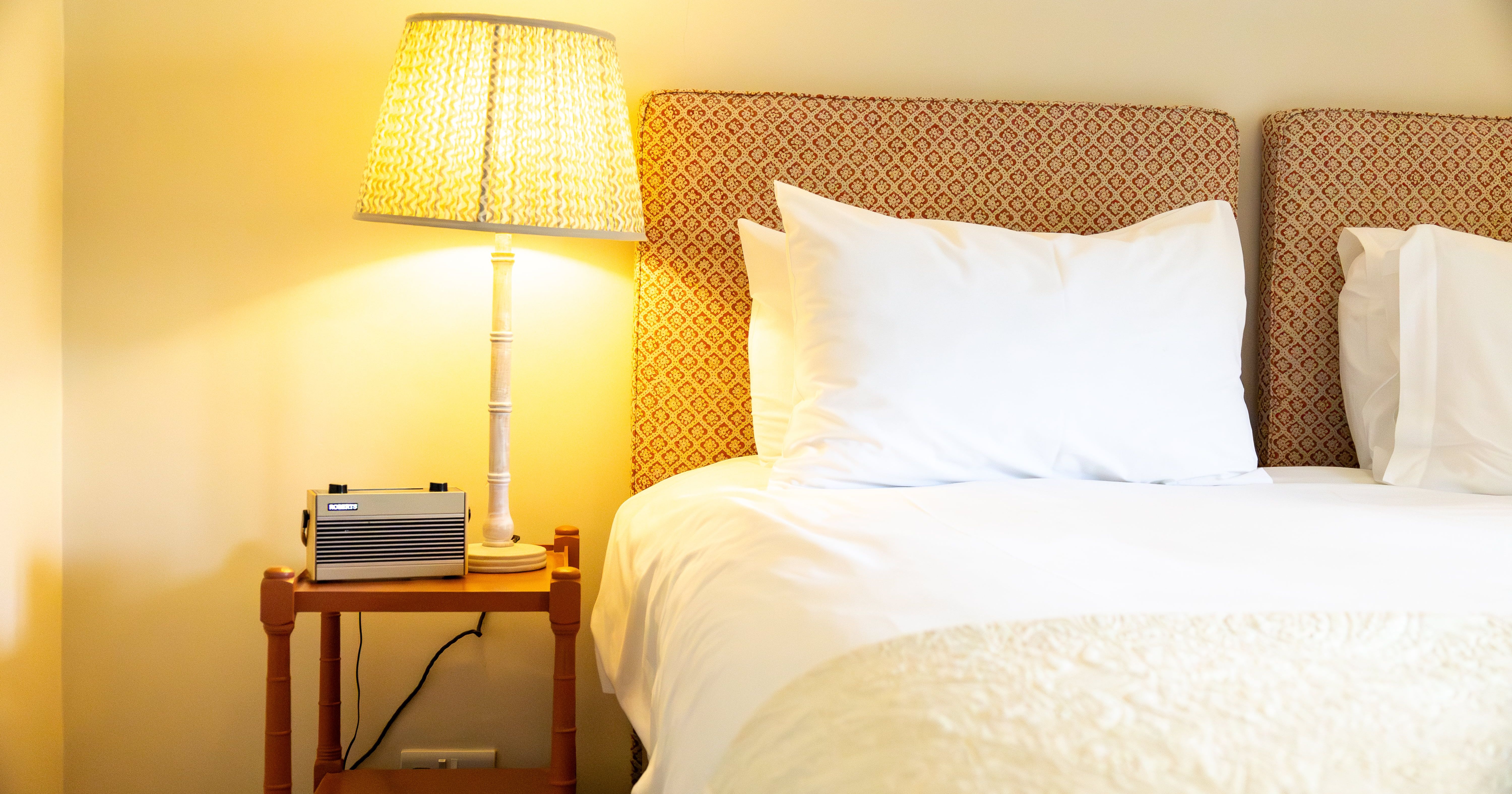 A cozy bedroom scene featuring a neatly made bed with white linens and several pillows, a patterned headboard, a wooden nightstand with a lamp, and a small retro radio.