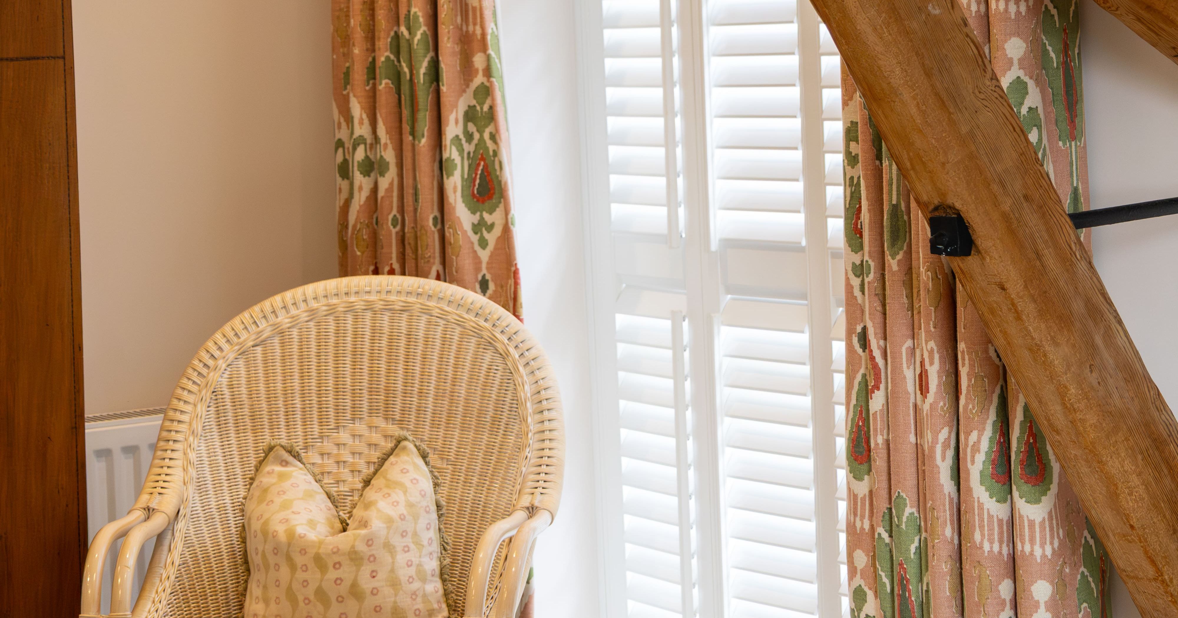 Wicker chair with cushion next to a window with patterned curtains and wooden beams