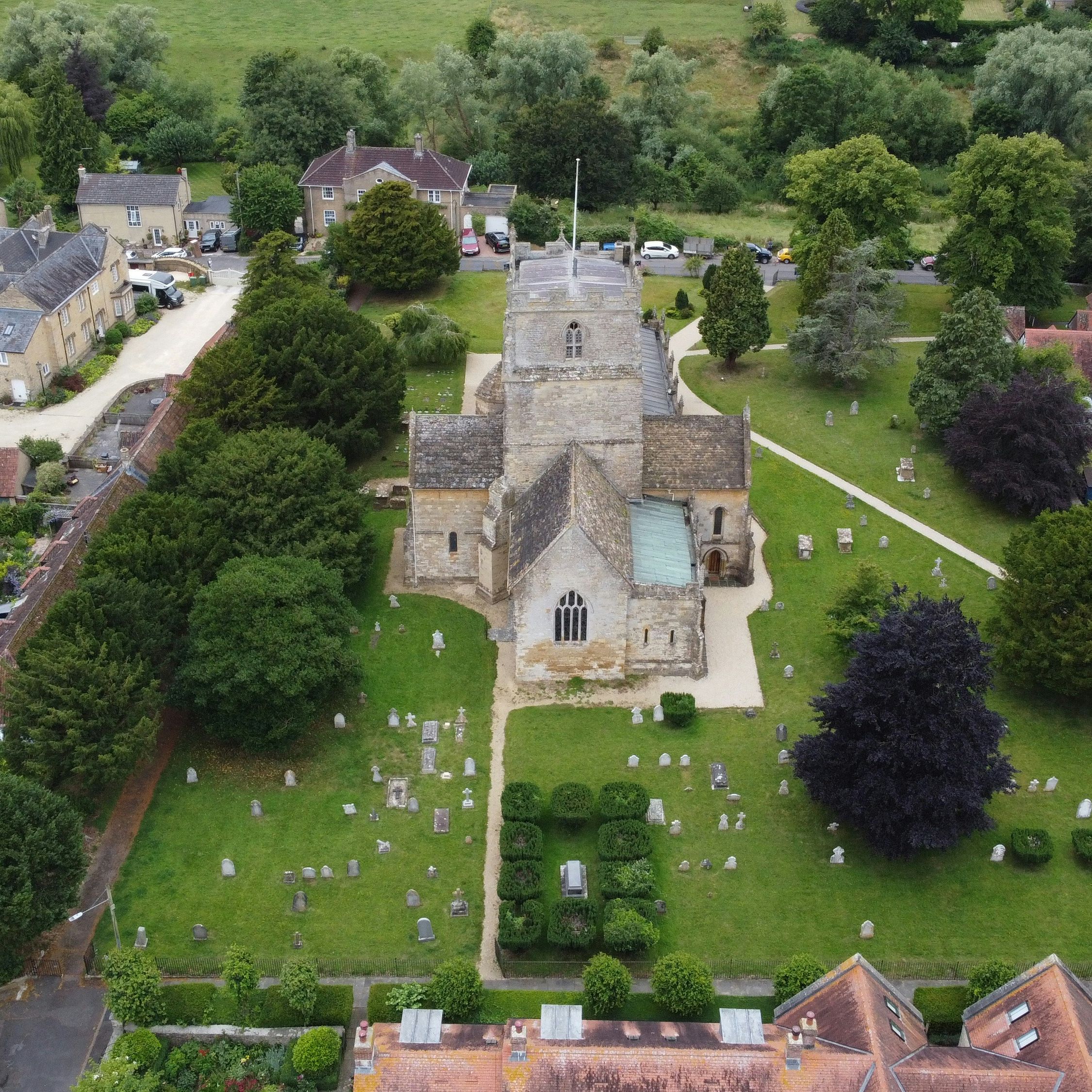 Aerial view of a historic stone church surrounded by a graveyard and trees in a rural village setting.