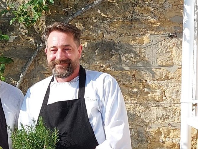 Two men in aprons standing outside a stone building, one holding vegetables and the other holding a potted herb.