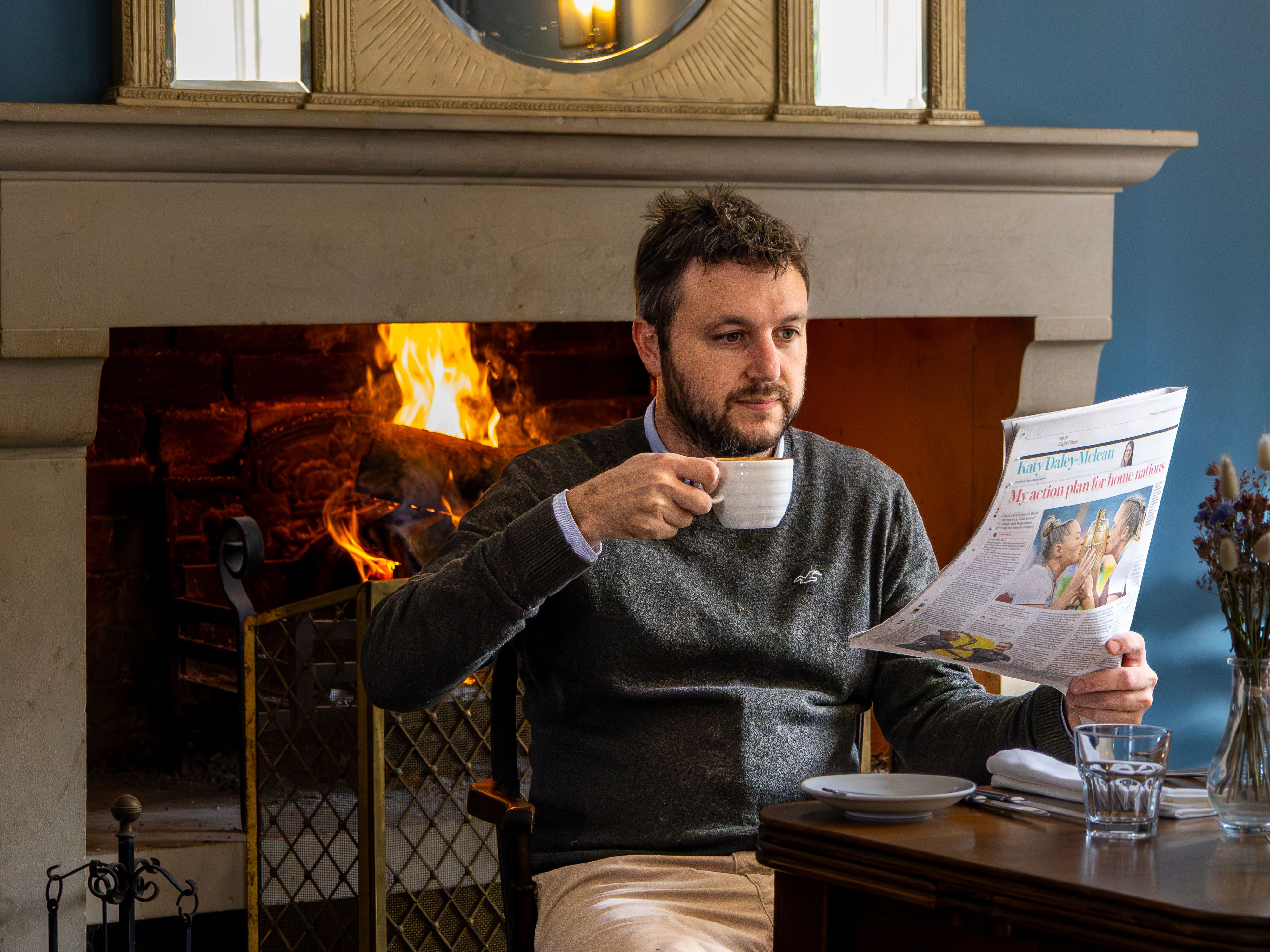Man sitting by a fireplace drinking coffee and reading a newspaper.