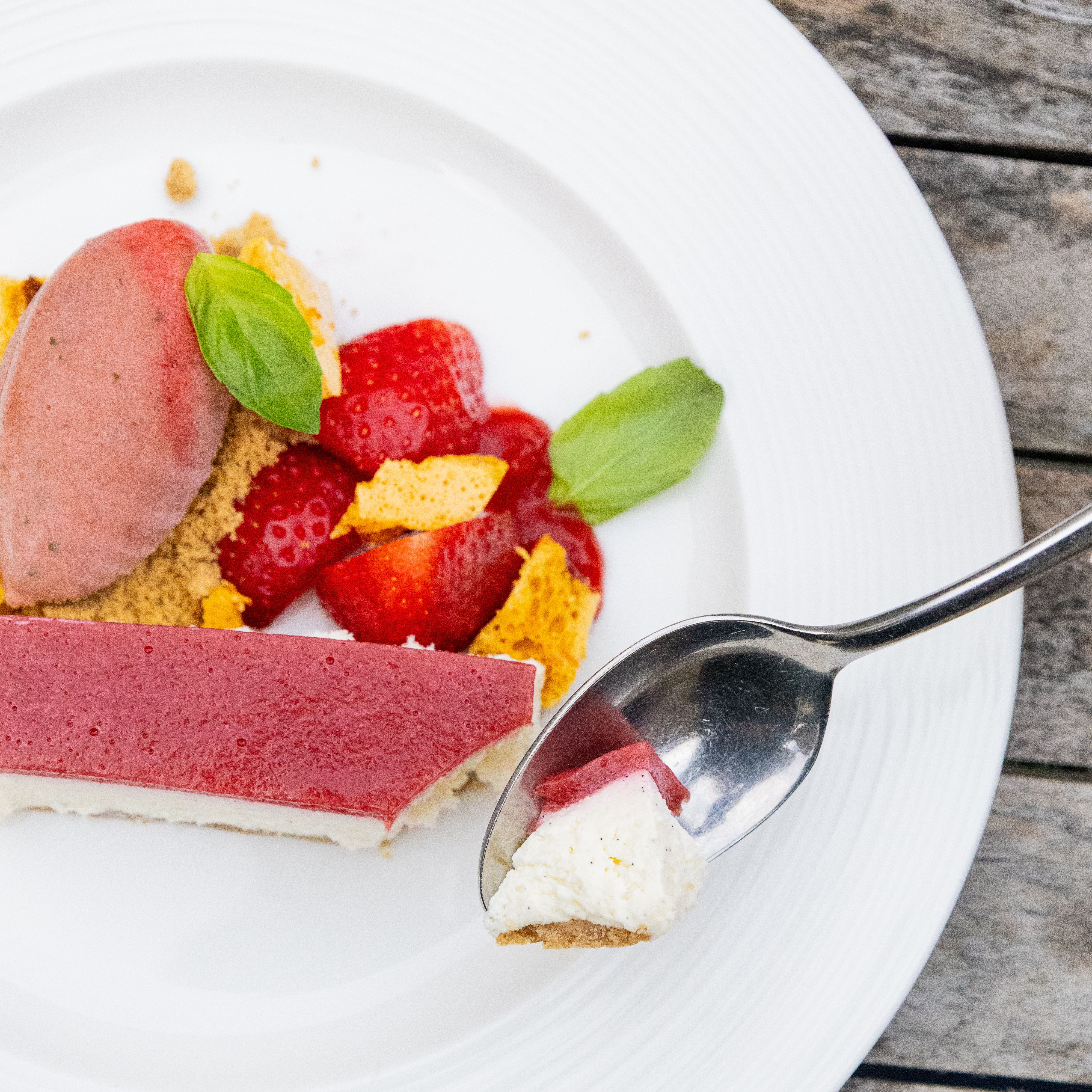 A plated dessert with strawberry cheesecake, fresh strawberries, strawberry sorbet, and a spoon taking a bite