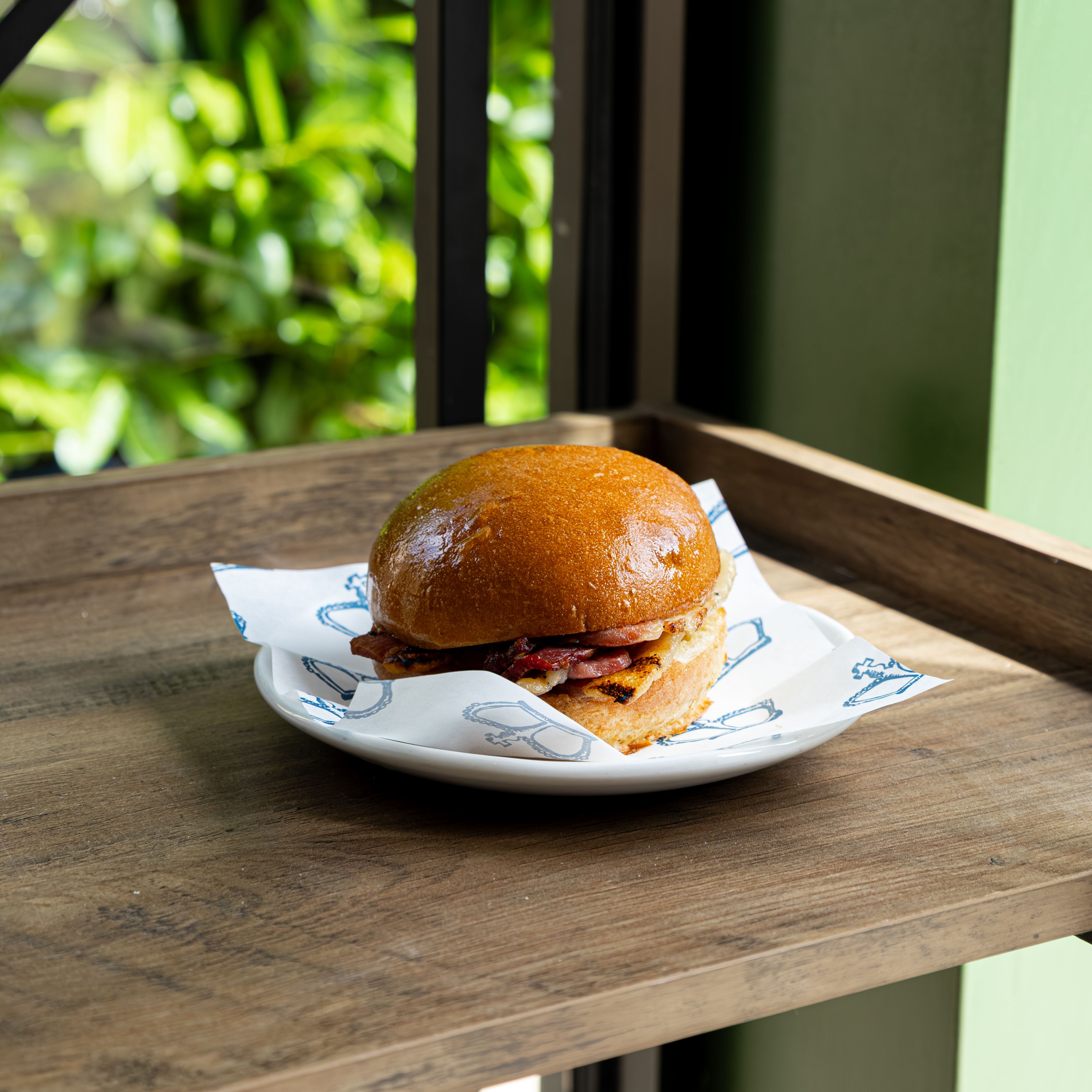 A bacon sandwich in a shiny brioche bun on a plate, placed on a wooden table by a window.