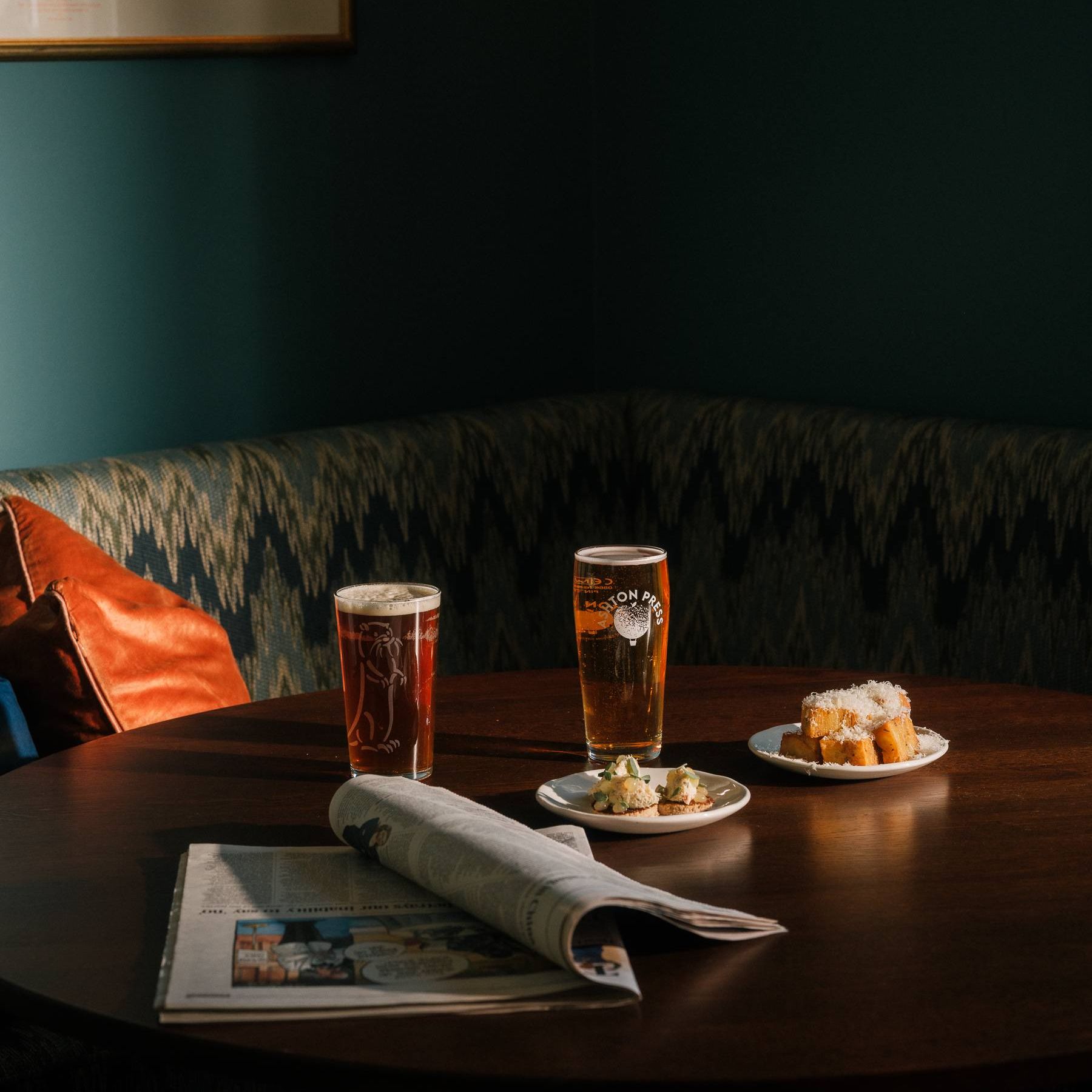 Two pints of beer and small plates on a pub table beside a newspaper in a cosy booth