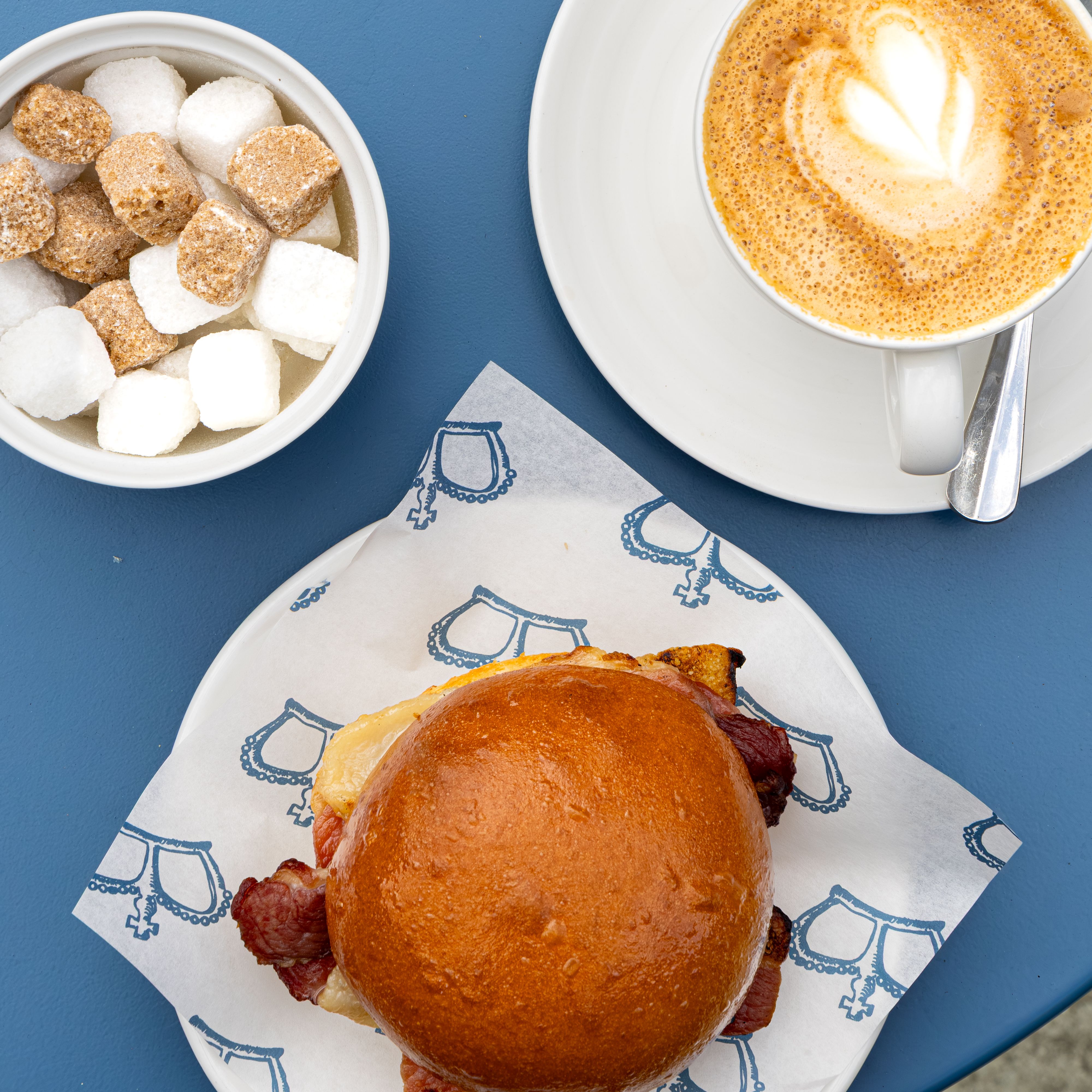 A cup of cappuccino, a bowl of sugar cubes, and a bacon sandwich on a blue table.