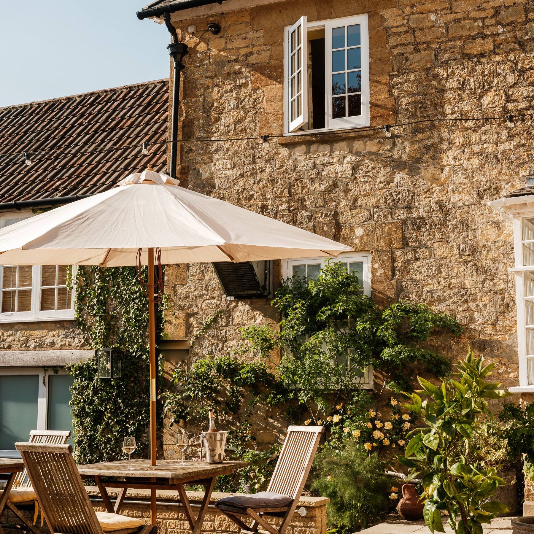 Sunlit stone cottage courtyard with wooden table and chairs under a white parasol